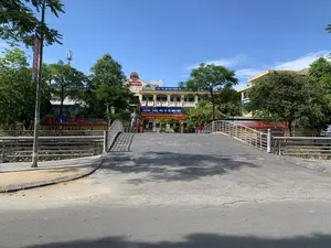 Bức ảnh chụp quang cảnh phía trước Trường Trung học cơ sở Điện Biên ở thành phố Thanh Hóa vào một ngày trời quang đãng. Tòa nhà trường học hai tầng, sơn màu vàng nhạt, có nhiều cửa sổ và cờ đỏ sao vàng được treo. Trên nóc tòa nhà có biển hiệu với logo và dòng chữ 'TRƯỜNG THCS ĐIỆN BIÊN'. Phía bên phải tòa nhà có một biểu ngữ màu xanh ghi '“VÌ LỢI ÍCH TRĂM NĂM...”'. Một cây cầu bộ hành nhỏ, cong, lát đá bắc qua một con kênh hoặc mương dẫn vào cổng trường. Một người đàn ông đang đi bộ trên cầu. Dọc theo lan can cầu và trước cổng trường có treo nhiều băng rôn màu đỏ. Một băng rôn lớn ở cổng ghi: 'TRƯỜNG TRUNG HỌC CƠ SỞ ĐIỆN BIÊN' và 'ĐẢNG BỘ, CHÍNH QUYỀN VÀ NHÂN DÂN PHƯỜNG ĐIỆN BIÊN QUYẾT TÂM XÂY DỰNG PHƯỜNG TRỞ THÀNH ĐƠN VỊ "ANH HÙNG LAO ĐỘNG TRONG THỜI KỲ ĐỔI MỚI"'. Địa chỉ trên băng rôn là 'ĐC: 99 TRIỆU QUỐC ĐẠT - P. ĐIỆN BIÊN - TP. THANH HÓA'. Các băng rôn khác có nội dung cảnh báo như '...NẠN ĐUỐI NƯỚC HỌC SINH KHÔNG ĐƯỢC RA SÁT MÉP HỒ' và 'KHÔNG ĐẬU XE - DỪNG XE TRÊN CẦU TRƯỚC CỔNG TRƯỜNG...'. Xung quanh có nhiều cây xanh và phía trước là một con đường nhựa. Phía xa có thể thấy một cột thu phát sóng viễn thông.。（日本語訳）。晴れた日の、タインホア市にあるディエンビエン中学校の正面の光景を写した写真です。2階建ての校舎は、明るい黄色に塗られ、多くの窓があり、金星紅旗が掲げられています。建物の屋上には、ロゴと「ディエンビエン中学校」という文字が入った看板があります。建物の右側には、「百年の利益のために…」と書かれた青い横断幕があります。石畳の小さなアーチ型の歩道橋が、運河または水路を渡って校門に通じています。一人の男性が橋の上を歩いています。橋の欄干沿いと校門の前に、多くの赤い横断幕が掲げられています。門にある大きな横断幕には、「ディエンビエン中学校」と「ディエンビエン区の党委員会、政府、人民は、区を『ドイモイ（刷新）期における労働英雄』単位に建設することを決意する」と書かれています。横断幕上の住所は「住所：99 TRIỆU QUỐC ĐẠT - ディエンビエン区 - タインホア市」です。他の横断幕には、「...生徒の溺水事故、池の縁に近づかないこと」や「校門前の橋の上での駐車・停車禁止...」のような警告内容があります。周囲には多くの緑の木々があり、手前にはアスファルトの道があります。遠くには、通信用の送受信塔が見えます。