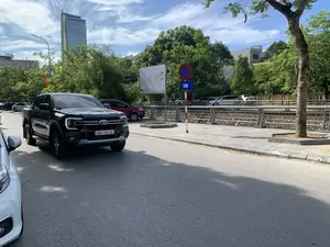 Quang cảnh một con đường đô thị vào một ngày nắng. Ở tiền cảnh, một chiếc xe bán tải màu đen hiệu Ford đang di chuyển trên đường nhựa, bên cạnh là một phần của chiếc ô tô màu trắng. Dọc bên đường có vỉa hè, lan can và nhiều cây xanh. Có một biển báo giao thông cấm dừng và đỗ xe, với biển phụ màu xanh ghi chữ '20M'. Ở phía sau, có nhiều ô tô khác đang đỗ, một tòa nhà cao tầng hiện đại bằng kính và các tòa nhà khác. Bầu trời trong xanh và có mây. Có một biển quảng cáo lớn màu trắng nhưng nội dung không đọc rõ và một biển hiệu có chữ 'Coffee' trên một tòa nhà ở phía xa.。（日本語訳）。晴れた日の都市の道路の光景。道沿いには歩道、ガードレール、そして多くの緑の木々がある。停車および駐車を禁止する交通標識があり、「20M」と書かれた青い補助標識が付いている。後方には、駐車している他の多くの自動車、ガラス張りの近代的な高層ビル、そしてその他の建物がある。空は澄んだ青色で、雲がある。大きな白い広告看板があるが、内容ははっきりと読み取れない。また、遠くの建物には「Coffee」という文字の看板がある。