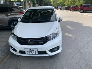 Bức ảnh chụp một chiếc ô tô Honda Brio RS màu trắng đang đỗ ven một con phố trải nhựa. Trên lưới tản nhiệt của xe có logo 'RS'. Khung cảnh xung quanh là một con đường đô thị với cây xanh hai bên. Nhiều ô tô khác cũng đang đỗ dọc theo con đường, và ở phía xa có hai người đang đi xe máy. Bên lề đường, có thể thấy một phần biển hiệu của ngân hàng 'VietinBank' và một biển báo tên đường ghi 'PHỐ LÊ QUÝ...', phần còn lại không thể đọc rõ. Trên kính chắn gió của chiếc xe màu trắng có dán một tem ghi số điện thoại '0962183888'.。（日本語訳）。写真は、舗装された通りの脇に駐車している白いホンダ・ブリオRSを写したものです。車のフロントグリルには「RS」のロゴがあります。周囲の風景は、両側に緑の木々が植えられた都市の道路です。他の多くの車も道路に沿って駐車しており、遠くにはバイクに乗った二人がいます。道端には、「VietinBank」銀行の看板の一部と、「PHỐ LÊ QUÝ...」（レ・クイ通り...）と書かれた通りの名前の標識が見えますが、残りの部分ははっきりと読み取れません。白い車のフロントガラスには、電話番号「0962183888」が記載されたステッカーが貼られています。