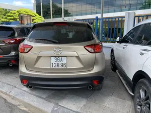 Bức ảnh chụp phía sau của một chiếc ô tô Mazda CX-5 màu vàng cát đang đỗ trong một bãi đỗ xe. Bãi đỗ xe được lát gạch. Ở phía sau là một tòa nhà hiện đại có mặt tiền bằng kính, được ngăn cách bởi một hàng rào kim loại màu xanh lam. Bên cạnh chiếc xe Mazda là một chiếc ô tô màu trắng ở bên phải và một chiếc ô tô màu xám đậm ở bên trái. Cảnh vật tĩnh, cho thấy các phương tiện đang được đỗ. Trên thân xe có các dòng chữ có thể đọc được là 'CX-5' và 'SKYACTIV TECHNOLOGY'.。（日本語訳）。駐車場に停まっている砂色のマツダCX-5の後ろ姿を写した写真です。駐車場はタイルで舗装されています。背景にはガラス張りの近代的な建物があり、青い金属製のフェンスで仕切られています。マツダ車の隣には、右側に白い車、左側に濃い灰色の車があります。静かな光景で、車両が駐車されていることがわかります。車体には「CX-5」と「SKYACTIV TECHNOLOGY」という文字が読み取れます。
