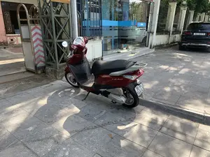 Hình ảnh ghi lại cảnh trên vỉa hè trước một tòa nhà. Ở tiền cảnh, một chiếc xe máy tay ga hiệu Honda Lead màu đỏ đang đỗ. Phía sau xe máy là một tòa nhà có cửa kính lớn, bên trong có một cây ATM và một người đang thực hiện giao dịch. Trên cửa kính có chữ "ATM". Phía trên có một biển hiệu màu vàng với dòng chữ "... Quốc Đạt" nhưng không nhìn rõ toàn bộ. Bên trái là một cánh cổng sắt dạng lùa có bánh xe, trên đó có các vạch cảnh báo chéo màu đỏ và trắng. Ở phía bên phải, một phần của chiếc ô tô màu đen đang đỗ. Vỉa hè được lát bằng gạch vuông màu xám, và có bóng nắng in trên mặt đất.。（日本語訳）。ある建物の前の歩道での光景を記録した画像です。前景には、ホンダ・リードブランドの赤いスクーターが1台駐車しています。スクーターの後ろには大きなガラス戸のある建物があり、中にはATMが1台と、取引を行っている人が一人います。ガラス戸には「ATM」という文字があります。その上には「... Quốc Đạt」という文字が入った黄色い看板がありますが、全体ははっきりと見えません。左側には車輪付きの引き戸式の鉄の門があり、その上には赤と白の斜めの警告縞模様があります。右側には、駐車している黒い車の一部が見えます。歩道は灰色の四角いタイルで舗装されており、地面には日差しによる影が映っています。