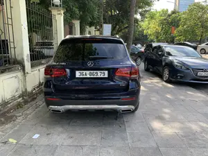 Bối cảnh là một vỉa hè trên một con phố đô thị. Một chiếc ô tô Mercedes-Benz GLC 200 4MATIC màu xanh đậm đang đỗ trên vỉa hè lát gạch đá. Trên đuôi xe có các chữ "GLC 200" và "4MATIC". Bên cạnh là một chiếc ô tô Mazda màu xanh đậm và các phương tiện khác cũng đang đỗ. Dọc theo vỉa hè là hàng rào sắt với các trụ bê tông và nhiều cây xanh. Ở phía sau, có một biển báo màu xanh dương treo trên cột điện với dòng chữ có thể đọc được là "ĐOÀN PHƯỜNG", "THANH NIÊN", "TỰ QUẢN". Xa hơn là các tòa nhà và cây cối khác.。（日本語訳）。背景は、都市の通りにある歩道です。濃い青色のメルセデス・ベンツGLC 200 4MATICが、石畳の歩道に駐車しています。車の後部には「GLC 200」と「4MATIC」の文字があります。隣には濃い青色のマツダ車と、他の車両も駐車しています。歩道に沿って、コンクリートの柱がある鉄のフェンスと多くの緑の木々があります。後方には、電柱に青い看板が掲げられており、「ĐOÀN PHƯỜNG」、「THANH NIÊN」、「TỰ QUẢN」という文字が読み取れます。さらに遠くには、他の建物や木々があります。