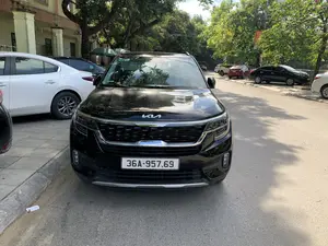 Bức ảnh chụp một chiếc ô tô màu đen hiệu KIA đang đỗ ở lề một con đường trong đô thị. Con đường được trải nhựa và có nhiều cây xanh lớn ở hai bên, tạo bóng mát xuống mặt đường. Ngoài chiếc xe KIA ở tiền cảnh, có nhiều xe ô tô khác (màu trắng, đen, đỏ) cũng đang đỗ dọc theo vỉa hè. Bên trái là một tòa nhà có các cột trụ và hàng rào sắt. Trên mui xe có logo chữ 'KIA'. Ở phía xa có thể thấy các biển hiệu và một băng rôn màu đỏ nhưng không thể đọc rõ nội dung. Khung cảnh yên tĩnh, không có người qua lại.。（日本語訳）。都市部の道路の脇に駐車しているKIAブランドの黒い自動車を写した写真です。道路はアスファルトで舗装されており、両側には大きな緑の木々が多く、路面に木陰を作っています。前景にあるKIAの車の他に、他の多くの自動車（白、黒、赤）も歩道に沿って駐車しています。左側には、柱と鉄柵のある建物があります。ボンネットには「KIA」の文字のロゴがあります。遠くには看板と赤い横断幕が見えますが、内容ははっきりと読み取れません。静かな光景で、通行人はいません。