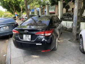 Bối cảnh là một con đường đô thị có vỉa hè lát gạch. Một chiếc ô tô Toyota Vios màu đen, bám bụi, đang đỗ trên vỉa hè. Trên cản sau của xe có một nhãn dán ghi 'TOYOTA DOANH THU - THANH HÓA'. Phía trước chiếc Vios là một chiếc ô tô Mazda màu xám. Cả hai chiếc xe đều mang biển kiểm soát của tỉnh Thanh Hóa (đầu số 36A). Dọc vỉa hè có trồng nhiều cây xanh. Có một cột điện bê tông, trên đó có ghi một dãy số bằng sơn màu xanh: '0915186 816'. Ở phía xa, có thể thấy một biển báo giao thông cấm rẽ trái. Khung cảnh cho thấy các phương tiện đang đỗ, không có hoạt động cụ thể nào của con người diễn ra ở cận cảnh.。（日本語訳）。背景は、レンガが敷かれた歩道のある都市の道路です。黒色で埃をかぶったトヨタ・ヴィオスが歩道に駐車しています。車の後部バンパーには「TOYOTA DOANH THU - THANH HÓA」と書かれたステッカーが貼られています。ヴィオスの前には灰色のマツダ車があります。2台ともタインホア省のナンバープレート（プレート番号の先頭が36A）を付けています。歩道沿いには多くの緑の木が植えられています。コンクリートの電柱があり、そこには青いペンキで「0915186 816」という数字が書かれています。遠くには、左折禁止の交通標識が見えます。この光景は車両が駐車していることを示しており、前景では特定の人の活動は見られません。
