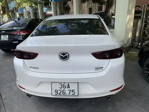 Hình ảnh chụp từ phía sau một chiếc ô tô màu trắng hiệu Mazda 3 đang đỗ trên vỉa hè lát gạch. Trên đuôi xe có các dòng chữ "MAZDA 3", "SKYACTIV G" và "DELUXE". Bên cạnh chiếc xe màu trắng là một chiếc ô tô khác màu đen, hiệu Toyota. Cả hai xe đều có biển số đăng ký tại tỉnh Thanh Hóa (mã vùng 36). Bối cảnh phía sau là một hàng rào sắt, cây xanh và một phần của một tòa nhà. Có một biển báo màu xanh ở phía xa nhưng không thể đọc rõ nội dung.。（日本語訳）。タイル敷きの歩道に駐車している白いマツダ3の後ろから撮影した画像。車の後部には「MAZDA 3」、「SKYACTIV G」、「DELUXE」の文字がある。白い車の隣には、もう一台の黒いトヨタ車がある。両方の車はタインホア省（地域コード36）で登録されたナンバープレートを付けている。遠くに青い看板があるが、内容ははっきりと読み取れない。