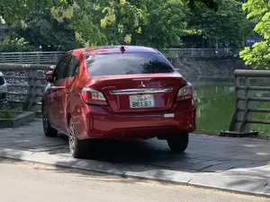 Trong ảnh là một chiếc ô tô hiệu Mitsubishi Attrage màu đỏ đang đỗ trên vỉa hè lát gạch bên cạnh một hồ nước. Phía sau xe là mặt hồ phẳng lặng và lan can bảo vệ. Bối cảnh phía xa là một cây cầu hoặc đường đi bộ ven hồ, với rất nhiều cây xanh. Nổi bật là những chùm hoa màu vàng, có thể là hoa muồng hoàng yến, đang nở rộ. Có thể nhìn thấy một biển báo chỉ dẫn bãi đỗ xe (biển báo chữ P) và một tấm biển lớn màu xanh dương ghi chữ "HỒ THA...", có khả năng là biển tên của hồ. Trên đuôi xe có chữ "Attrage". Khung cảnh chung là một không gian công cộng yên tĩnh ven hồ tại một đô thị.。（日本語訳）。車の後方には、穏やかな湖面と保護用の手すりがあります。遠景は橋または湖畔の遊歩道で、たくさんの緑樹があります。目立っているのは黄色い花の房で、おそらく満開のゴールデンシャワー（ナンバンサイカチ）の花でしょう。駐車場を示す標識（Pの文字の標識）と、「HỒ THA...」と書かれた大きな青い看板が見え、これは湖の名前の看板である可能性があります。車の後部には「Attrage」という文字があります。全体的な光景は、ある都市の湖畔にある静かな公共空間です。