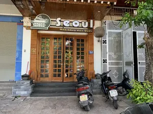 Hình ảnh chụp mặt tiền của một nhà hàng Hàn Quốc có tên là 'Seoul', địa chỉ tại số 152 đường Triệu Quốc Đạt. Biển hiệu của nhà hàng có chữ 'Seoul' và dòng chữ 'Nhà hàng Hàn Quốc'. Cửa ra vào chính được làm bằng gỗ và kính, hiện đang đóng. Phía trước nhà hàng, trên vỉa hè có hai chiếc xe máy đang đỗ. Bên phải lối vào chính là một cổng sắt màu trắng đang mở hé, và có một cái cây xanh. Một chiếc đèn lồng trang trí bằng tre đan được treo dưới mái hiên. Bên trái là một mặt bằng khác với cửa cuốn đang đóng. Lối vào nhà hàng có các bậc tam cấp bằng đá. Có camera an ninh được lắp đặt trên tường.。（日本語訳）。「ソウル」という名前の韓国料理店の正面を撮影した写真で、住所はチエウ・クオック・ダット通り152番地です。店の看板には「ソウル」という文字と「韓国料理店」という文言があります。正面の出入り口は木とガラスでできており、現在は閉まっています。店の前、歩道には2台のバイクが停まっています。正面の入り口の右側には、少し開いている白い鉄の門があり、緑の木が1本あります。軒下には竹で編んだ装飾用の提灯が吊るされています。左側は、シャッターが閉まっている別の店舗です。店の入り口には石の階段があります。壁には防犯カメラが設置されています。