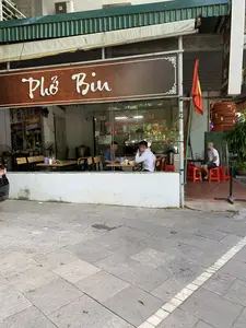 Trên biển hiệu chính của quán có ghi 'Phở Bin'. Trên cửa kính có dán thông tin thực đơn và giá cả, bao gồm 'Phở', 'Bún Phở', 'Tái - Bắp - Gầu - Nạm 35K', và 'Phở Đặc Biệt 50K'. Một tấm biển khác có vẻ ghi 'MIỄN PHÍ TRÀ ĐÁ TỰ PHỤC VỤ' (chữ 'MIEN TRA TỰ PH' có thể nhìn thấy).。Bối cảnh là một quán phở ven đường. Bên trong quán, qua lớp cửa kính, có thể thấy hai người đàn ông đang ngồi ăn tại bàn gỗ. Bên ngoài hiên, một người đàn ông khác đang ngồi trên ghế nhựa màu đỏ. Phía trước quán có treo một lá cờ Việt Nam (cờ đỏ sao vàng). Không gian quán có bàn ghế gỗ, quạt điện, và vỉa hè phía trước được lát gạch đá.。（日本語訳）。店のメインの看板には「Phở Bin」と書かれています。ガラスのドアには、「フォー」、「ブンフォー」、「タイ - バップ - ガウ - ナム 35K」、そして「特別フォー 50K」を含むメニューと価格の情報が貼られています。別の看板には「MIỄN PHÍ TRÀ ĐÁ TỰ PHỤC VỤ」（無料アイスティーセルフサービス）と書かれているようです（「MIEN TRA TỰ PH」の文字が見えます）。背景は道端のフォーの店です。店内では、ガラスのドア越しに、2人の男性が木製のテーブルで食事をしているのが見えます。軒先では、別の男性が赤いプラスチックの椅子に座っています。店の前にはベトナムの国旗（金星紅旗）が掲げられています。店内には木製のテーブルと椅子、扇風機があり、店の前の歩道は石のタイルで舗装されています。
