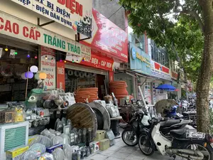 Bản ghi văn bản từ các biển hiệu:。1. Cửa hàng 'CAO CẤP TRÁNG THẢO' (Lô 20 Triệu Quốc Đạt, ĐT: 0373.855.458), là đại lý thiết bị điện và cáp điện, quảng cáo các thương hiệu CADI-SUN, CADI-GOLD, GOLDHAN.。2. 'CÔNG TY TNHH DŨNG XUÂN' (Lô 19 Triệu Quốc Đạt, Phường Điện Biên, TP Thanh Hóa, SĐT: 0902 225 365), chuyên về thiết bị điện, điện tự động hóa và điện công nghiệp.。3. 'Niêu Quán' (334 Đường Triệu Quốc Đạt, TP. Thanh Hóa), phục vụ cơm niêu, cơm viên phố và cà phê sách.。4. 'Vip Mart' (Lô 16-17 Triệu Quốc Đạt, TP. Thanh Hóa), bán hàng cho mẹ và bé và hàng tiêu dùng nhập khẩu.。Mô tả cảnh quan và hoạt động:。Bức ảnh chụp một dãy cửa hàng mặt tiền trên một con phố ở thành phố Thanh Hóa. Cửa hàng nổi bật nhất là 'Tráng Thảo', một cửa hàng bán thiết bị điện. Hàng hóa được trưng bày tràn ra vỉa hè, bao gồm các cuộn cáp điện lớn trên lõi gỗ, các cuộn ống luồn dây điện màu cam, nhiều loại quạt điện, đèn chiếu sáng và các thiết bị điện khác. Các cửa hàng đang mở cửa kinh doanh. Nhiều xe máy (xe gắn máy) đang được đỗ trên vỉa hè phía trước các cửa hàng, và có một cây xanh lớn ở bên phải.。（日本語訳）。看板からのテキスト記録：。1. 店舗「CAO CẤP TRÁNG THẢO」（ロット20、チウ・クオック・ダット通り、電話：0373.855.458）は、電気設備および電線ケーブルの代理店で、CADI-SUN、CADI-GOLD、GOLDHANのブランドを広告しています。2. 「DŨNG XUÂN有限会社」（ロット19、チウ・クオック・ダット通り、ディエンビエン区、タインホア市、電話番号：0902 225 365）は、電気設備、電気オートメーション、産業用電気を専門としています。3. 「Niêu Quán」（334 チウ・クオック・ダット通り、タインホア市）は、土鍋ご飯、ストリート風ライスボール、ブックカフェを提供しています。4. 「Vip Mart」（ロット16-17、チウ・クオック・ダット通り、タインホア市）は、マタニティ・ベビー用品および輸入消費財を販売しています。風景と活動の描写：。写真はタインホア市のある通りに面した店舗の列を撮影したものです。最も目立つ店舗は、電気設備を販売する「Tráng Thảo」です。商品は歩道にあふれるように陳列されており、木製の芯に巻かれた大きな電線ケーブルの束、オレンジ色の電線管の束、様々な種類の扇風機、照明器具、その他の電気設備が含まれています。各店舗は営業中です。多くのバイク（原動機付自転車）が店舗前の歩道に駐車されており、右側には大きな緑の木があります。