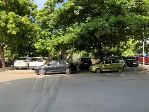 Hình ảnh chụp một bãi đỗ xe ven đường vào một ngày nắng. Nhiều chiếc ô tô, bao gồm các loại xe SUV, sedan và hatchback với các màu sắc khác nhau như trắng, đen, và xanh lục, đang đỗ trên vỉa hè dưới bóng mát của những hàng cây cổ thụ xanh tốt. Tán lá rậm rạp của cây che phủ phần lớn khu vực, tạo ra những vệt bóng trên mặt đường nhựa. Ở phía sau, có thể thấy một cột sơn sọc đỏ trắng và một biển báo giao thông hình tròn màu xanh. Khung cảnh yên tĩnh, không có sự xuất hiện của người.。（日本語訳）。ある晴れた日の道端の駐車場を撮影した写真。白、黒、緑など様々な色のSUV、セダン、ハッチバックを含む多くの車が、青々と茂る古い並木道の木陰の歩道に駐車している。木々の鬱蒼とした葉がエリアの大部分を覆い、アスファルトの路面に影の筋を作っている。静かな光景で、人の姿はない。