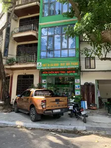 Bản ghi chép các thông tin văn bản trên hình ảnh:。Trên biển hiệu chính màu xanh lá cây: 'CÔNG TY CỔ PHẦN VINATECH', 'THIẾT BỊ AN NINH - MÁY VĂN PHÒNG'. Địa chỉ: 'ĐỊA CHỈ: 04 HÀNG ĐỒNG - TPTH', 'ĐT: 02373 75 75 78 - DĐ: 0982 998 221 WEB: VinaTech.Vnn.Vn'.。Trên bảng đèn LED màu đỏ: 'SỬA CHỮA MÁY TÍNH - MÁY IN', 'CAMERA - BẢO ĐỘNG - CHẤM CÔNG - ĐẾM TIỀN'.。Trên decal dán ở đuôi xe bán tải: 'BÁN - CHO THUÊ MÁY PHOTOCOPY - MÁY IN' cùng với hai số điện thoại '0912.234.698 - 0911.266.386'.。Trên các biển quảng cáo nhỏ hơn bên trong và ngoài cửa hàng: 'CAMERA', 'BÁO ĐỘNG', 'KHÓA VÂN TAY', 'MÁY TÍNH', 'THIẾT BỊ MẠNG', 'vinaphone'.。Mô tả cảnh vật và hoạt động:。Đây là hình ảnh chụp mặt tiền một cửa hàng kinh doanh trên một con phố. Địa điểm là một tòa nhà nhiều tầng, trong đó tầng trệt là cửa hàng 'Công ty Cổ phần Vinatech' với mặt tiền được sơn màu xanh lá cây và có cửa kính lớn. Cửa hàng này chuyên kinh doanh và sửa chữa thiết bị an ninh (camera, báo động) và máy văn phòng (máy tính, máy in, máy chấm công, máy đếm tiền).。Phía trước cửa hàng, trên vỉa hè, có một chiếc xe bán tải hiệu Ford Ranger màu cam và hai chiếc xe máy đang đỗ. Bên cạnh là một cái cây lớn, một tủ gỗ, một máy nén khí và một biển quảng cáo của Vinaphone. Có một vài người dường như là nhân viên hoặc khách hàng đang ở bên trong cửa hàng.。（日本語訳）。画像上のテキスト情報の記録：。緑色のメイン看板には、「VINATECH株式会社」、「セキュリティ機器 - オフィス機器」。住所：「住所：04 HANG DONG - TPTH」、「電話：02373 75 75 78 - 携帯：0982 998 221 WEB：VinaTech.Vnn.Vn」。赤色のLED電光掲示板には、「コンピューター - プリンター修理」、「カメラ - 警報 - 勤怠管理 - 紙幣計数機」。ピックアップトラックの後部に貼られたデカールには、「コピー機 - プリンターの販売・レンタル」と2つの電話番号「0912.234.698 - 0911.266.386」。店舗の内外にある小さな広告看板には、「カメラ」、「警報」、「指紋認証ロック」、「コンピューター」、「ネットワーク機器」、「vinaphone」。風景と活動の説明：。これは、ある通りにある店舗の正面を撮影した画像です。場所は多層階の建物で、その1階が「Vinatech株式会社」の店舗となっており、正面は緑色に塗られ、大きなガラスのドアがあります。この店舗は、セキュリティ機器（カメラ、警報）とオフィス機器（コンピューター、プリンター、勤怠管理機、紙幣計数機）の販売と修理を専門としています。店舗の前、歩道には、オレンジ色のフォード・レンジャーのピックアップトラック1台とバイク2台が駐車しています。その隣には、大きな木、木製の棚、エアコンプレッサー、そしてVinaphoneの広告看板があります。店舗の中には、従業員か客と思われる数人の人がいます。