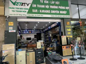Bức ảnh chụp mặt tiền và không gian bên trong của một cửa hàng kinh doanh thiết bị âm thanh tại thành phố Thanh Hóa.。**Thông tin văn bản:**。Trên tấm biển hiệu lớn màu xanh lá cây có các dòng chữ: "VIETKTV", "TRUNG TÂM BẢO HÀNH TẠI THANH HÓA", "TƯ VẤN - LẮP ĐẶT ÂM THANH HỘI TRƯỜNG PHÒNG HỌP BAR - KARAOKE CHUYÊN NGHIỆP". Địa chỉ được ghi rõ là "Add: 32+34 Hàng Đồng - P.Điện Biên - Tp.Thanh Hóa" và số điện thoại "Hotline: 0912.602.662 - 02373.753.335". Có hai biển số nhà ghi "32 HÀNG ĐỒNG" và "34 HÀNG ĐỒNG". Bên trong cửa hàng có biển hiệu "CÔNG TY TNHH ĐIỆN TỬ HẢI VÂN" cùng logo của các thương hiệu như JBL, SHURE, dbx.。**Miêu tả cảnh vật và hoạt động:**。*   **Loại địa điểm:** Đây là một cửa hàng điện tử, chuyên về các thiết bị âm thanh chuyên nghiệp và karaoke.。*   **Vật thể:** Cửa hàng trưng bày rất nhiều loại loa thùng (loa), amply, mixer và các thiết bị âm thanh khác. Nhiều loa có kích thước lớn, được xếp chồng lên nhau ở cả phía trước và bên trong, một số vẫn còn bọc trong lớp nilon. Ngoài ra còn có một vài chiếc quạt cây, một phần của một chiếc xe máy đậu bên trái, và các camera an ninh được lắp đặt bên ngoài.。*   **Hoạt động:** Một người đàn ông mặc áo đen đang ngồi xổm, tập trung vào việc kiểm tra hoặc sửa chữa một mạch điện tử của một thiết bị âm thanh. Một người khác đang ngồi ở phía sâu bên trong cửa hàng. Khung cảnh cho thấy cửa hàng đang trong giờ hoạt động kinh doanh và sửa chữa.。（日本語訳）。タインホア市にある音響機器を販売する店舗の正面と内部空間を撮影した写真。**テキスト情報：**。大きな緑色の看板には、「VIETKTV」、「TRUNG TÂM BẢO HÀNH TẠI THANH HÓA」（タインホア保証センター）、「TƯ VẤN - LẮP ĐẶT ÂM THANH HỘI TRƯỜNG PHÒNG HỌP BAR - KARAOKE CHUYÊN NGHIỆP」（コンサルティング - ホール・会議室・バー・プロフェッショナルカラオケの音響設置）という文言があります。住所は「Add: 32+34 Hàng Đồng - P.Điện Biên - Tp.Thanh Hóa」、電話番号は「Hotline: 0912.602.662 - 02373.753.335」と明記されています。「32 HÀNG ĐỒNG」と「34 HÀNG ĐỒNG」と書かれた2つの番地表示があります。店内には「CÔNG TY TNHH ĐIỆN TỬ HẢI VÂN」（ハイヴァン電子有限会社）という看板と、JBL、SHURE、dbxなどのブランドロゴがあります。**風景と活動の描写：**。*   **場所の種類：** これは電子機器店で、プロ仕様の音響機器とカラオケ機器を専門としています。*   **物体：** 店内には、多くの種類のスピーカーボックス（スピーカー）、アンプ、ミキサー、その他の音響機器が展示されています。多くのスピーカーは大型で、店の前と内部の両方で積み重ねられており、一部はまだナイロンで包まれています。その他、数台の扇風機、左側に駐車しているバイクの一部、そして外に設置された防犯カメラがあります。*   **活動：** 黒い服を着た男性がしゃがみ込み、音響機器の電子回路の点検または修理に集中しています。もう一人の人物が店の奥に座っています。この光景は、店が営業および修理の時間内であることを示しています。