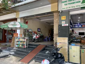 Bối cảnh là mặt tiền của hai cửa hàng kinh doanh nằm liền kề nhau trên một con phố ở Thanh Hóa. Cửa hàng bên trái là một tiệm tạp hóa nhỏ, trong khi cửa hàng bên phải chuyên bán thiết bị âm thanh. Phía trước cửa hàng bên trái có một chiếc ô lớn màu xanh lá cây với dòng chữ 'CHANH MUỐI 360' và một tấm biển màu đỏ ghi 'BÁN THẺ ĐIỆN THOẠI'. Bên trong tiệm tạp hóa có các kệ hàng, hai chiếc xe máy đang đậu và một người đang ngồi trên ghế nhựa. Cửa hàng bên phải có một biển hiệu lớn màu xanh lá cây ghi 'TRUNG TÂM BẢO... TẠI THANH HÓA' và địa chỉ 'Add: 32+34 Hàng Đồng...'. Một biển số nhà màu vàng ghi '32 HÀNG ĐỒNG'. Cửa hàng này trưng bày nhiều loa thùng lớn, một số vẫn còn bọc nilon, cùng với các thiết bị âm thanh khác. Các thương hiệu như NANOMAX, JBL, CROWN có thể được nhìn thấy. Một chiếc xe máy màu đen đang đỗ ở phía trước. Hoạt động chung là cảnh buôn bán thường ngày của các cửa hàng.。（日本語訳）。背景は、タインホア省のある通りに隣接して並ぶ2つの商店の正面です。左側の店は小さな雑貨店で、右側の店は音響機器を専門に販売しています。左側の店の前には、「CHANH MUỐI 360」という文字が入った大きな緑色の傘と、「BÁN THẺ ĐIỆN THOẠI」（携帯電話カード販売）と書かれた赤い看板があります。雑貨店の中には、商品棚、駐車している2台のバイク、そしてプラスチックの椅子に座っている人が一人います。右側の店には、「TRUNG TÂM BẢO... TẠI THANH HÓA」（タインホアの...保証センター）と書かれた大きな緑色の看板と、「Add: 32+34 Hàng Đồng...」という住所があります。「32 HÀNG ĐỒNG」と書かれた黄色い番地プレートがあります。この店は、いくつかはまだナイロンで包まれたままの大きなスピーカーボックスを多数、他の音響機器と一緒に展示しています。NANOMAX、JBL、CROWNなどのブランドが見られます。一台の黒いバイクが前に停まっています。全体的な活動は、商店の日常的な商売の光景です。