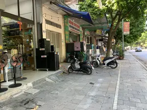 Bối cảnh là một con phố tại Việt Nam với các cửa hàng kinh doanh nằm dọc vỉa hè. Vỉa hè được lát đá màu xám và có cây xanh. Các cửa hàng đang mở cửa, trưng bày sản phẩm ra bên ngoài.。Các thông tin bằng chữ có thể nhận dạng:。- Biển hiệu cửa hàng màu xanh lá cây: "ĐIỆN MÁY THÀNH THANH", "MÁY PHÁT ĐIỆN CÔNG CỤ CẦM TAY", "CHO THUÊ MÁY PHÁT ĐIỆN", và số điện thoại "ĐT: 0913 529 568 - 0949 025 968".。- Số nhà: "36" và "38".。- Biểu ngữ màu đỏ treo trên cột: "LỰA CHỌN THỰC PHẨM RÕ NGUỒN GỐC XUẤT XỨ CHO BỮA ĂN NGON, AN TOÀN SỨC KHỎE" và logo "SAVANI".。- Tên thương hiệu trên sản phẩm: "HA VAN" trên loa, "JAGUAR" bên trong cửa hàng kính, "Tiger" trên các thùng các-tông.。Các vật thể và hoạt động:。- Địa điểm: Một con phố kinh doanh. Bên trái là cửa hàng điện tử, âm thanh với nhiều loa thùng lớn và quạt cây được bày bán. Bên cạnh là cửa hàng cho thuê và bán máy phát điện.。- Hoạt động: Cảnh đường phố ban ngày, không có hoạt động mua bán cụ thể nào đang diễn ra. Có xe cộ lưu thông trên đường.。- Vật thể: Nhiều loa thùng màu đen, quạt cây, máy phát điện công nghiệp, các chồng nồi inox lớn, hai chiếc xe máy (một chiếc màu đen hiệu SYM Elite 50 và một chiếc màu trắng) đang đỗ trên vỉa hè. Dưới lòng đường có một chiếc ô tô màu đen đang chạy.。（日本語訳）。背景はベトナムのある通りで、歩道に沿って商店が並んでいます。歩道は灰色の石で舗装されており、植木があります。店は営業しており、商品を外に陳列しています。認識可能な文字情報：。- 緑色の店の看板：「ĐIỆN MÁY THÀNH THANH」、「MÁY PHÁT ĐIỆN CÔNG CỤ CẦM TAY」（発電機・携帯工具）、「CHO THUÊ MÁY PHÁT ĐIỆN」（発電機レンタル）、および電話番号「ĐT: 0913 529 568 - 0949 025 968」。- 番地：「36」と「38」。- 柱に掲げられた赤い横断幕：「LỰA CHỌN THỰC PHẨM RÕ NGUỒN GỐC XUẤT XỨ CHO BỮA ĂN NGON, AN TOÀN SỨC KHỎE」（美味しく、健康に安全な食事のために、出所・由来が明確な食品を選びましょう）および「SAVANI」のロゴ。- 製品上のブランド名：スピーカーの「HA VAN」、ガラス張りの店内の「JAGUAR」、段ボール箱の「Tiger」。物体と活動：。- 場所：商店街。左側は電化製品・音響店で、多くの大型スピーカーと扇風機が販売のために陳列されています。その隣は発電機のレンタル・販売店です。- 活動：日中の街の風景で、特定の売買活動は行われていません。道路には車両が通行しています。- 物体：多くの黒いスピーカー、扇風機、業務用発電機、積み重ねられた大きなステンレス鍋、2台のバイク（1台は黒のSYM Elite 50、もう1台は白）が歩道に停まっています。車道には1台の黒い車が走っています。