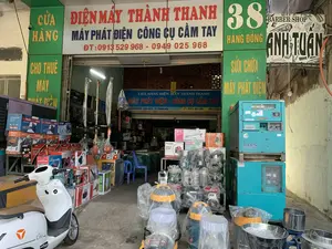 Đây là hình ảnh mặt tiền của một cửa hàng kinh doanh máy móc thiết bị tại địa chỉ 38 Hàng Đồng, Thành phố Thanh Hóa. Cửa hàng có tên là 'ĐIỆN MÁY THÀNH THANH', chuyên bán, cho thuê và sửa chữa máy phát điện và công cụ cầm tay.。Nội dung các biển hiệu có thể đọc được:。- Biển hiệu chính: 'ĐIỆN MÁY THÀNH THANH', 'MÁY PHÁT ĐIỆN CÔNG CỤ CẦM TAY', và số điện thoại 'ĐT: 0913 529 968 - 0949 025 968'.。- Các biển hiệu dọc màu xanh lá cây: 'CỬA HÀNG', 'CHO THUÊ MÁY PHÁT ĐIỆN', '38 HÀNG ĐỒNG', 'SỬA CHỮA MÁY PHÁT ĐIỆN'.。- Biển hiệu bên trong cửa hàng: 'CỬA HÀNG ĐIỆN MÁY THÀNH THANH', 'MÁY PHÁT ĐIỆN - CÔNG CỤ CẦM TAY', '38 HÀNG ĐỒNG - ĐIỆN BIÊN - TP. THANH HÓA'.。- Biển hiệu của cửa hàng bên cạnh: 'BARBER SHOP ANH TUẤN', '42 HÀNG ĐỒNG'.。Các vật thể và hoạt động trong ảnh:。- Địa điểm: Một cửa hàng điện máy mặt phố.。- Hoạt động: Trưng bày hàng hóa để bán và cho thuê. Không có người trong ảnh nhưng cửa hàng đang mở cửa kinh doanh.。- Vật thể: Rất nhiều loại máy móc được trưng bày, bao gồm các máy phát điện công nghiệp lớn màu xanh lam (hiệu Denyo), máy hút bụi công nghiệp, máy cắt cỏ, máy xay bằng inox, và nhiều hộp sản phẩm công cụ cầm tay như máy cưa xích, máy rửa xe áp lực cao, máy hàn. Một chiếc xe máy điện màu trắng (hiệu Orla) đang đỗ ở phía trước.。（日本語訳）。これは、タインホア市ハン・ドン通り38番地にある機械設備を扱う店の正面の写真です。店の名前は「ディエンマイ・タイン・タイン」で、発電機と手持ち工具の販売、レンタル、修理を専門としています。読み取れる看板の内容：。- メインの看板：「ディエンマイ・タイン・タイン」、「発電機 手持ち工具」、および電話番号「電話：0913 529 968 - 0949 025 968」。- 緑色の縦長の看板：「店舗」、「発電機レンタル」、「ハン・ドン通り38番」、「発電機修理」。- 店内の看板：「ディエンマイ・タイン・タイン店」、「発電機 - 手持ち工具」、「ハン・ドン通り38番 - ディエンビエン区 - タインホア市」。- 隣の店の看板：「バーバーショップ・アイン・トゥアン」、「ハン・ドン通り42番」。写真の中の物体と活動：。- 場所：通りに面した電化製品店。- 活動：販売およびレンタル用の商品を陳列している。写真には人が写っていないが、店は営業中である。- 物体：非常に多くの種類の機械が陳列されており、青色の大型産業用発電機（デンヨー製）、産業用掃除機、草刈り機、ステンレス製の粉砕機、そしてチェーンソー、高圧洗浄機、溶接機などの多くの手持ち工具の製品箱が含まれている。一台の白色の電動バイク（Orla製）が正面に停まっている。