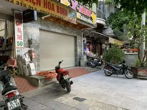 Khung cảnh chụp một con phố tại khu vực đô thị, với các cửa hàng kinh doanh nằm san sát nhau trên vỉa hè. Các biển hiệu cho thấy đây có thể là phố Hàng Đồng, chuyên bán các mặt hàng điện. Các dòng chữ trên biển hiệu có thể đọc được gồm: 'ĐIỆN CAO HẠ THẾ 50 HÀNG ĐỒNG', 'HÒA BÌNH' cùng số điện thoại '0932 312 313', 'X-MEN 52 HÀNG ĐỒNG', và 'Xuân Quang 54 Hàng Đồng'. Một cửa hàng có cửa cuốn đang đóng. Hoạt động chính là một người đàn ông lớn tuổi đang ngồi trên ghế nhựa và sử dụng điện thoại di động. Xung quanh ông là các mặt hàng điện như đèn chiếu sáng còn trong hộp và một cột thu lôi. Trên vỉa hè có hai chiếc xe máy đang đỗ, cùng với các cuộn ống luồn dây điện màu cam. Gần đó có một tủ điện với biển cảnh báo 'CẤM LẠI GẦN。CÓ ĐIỆN NGUY HIỂM CHẾT NGƯỜI'. Cảnh quan còn có cây xanh và một chậu cây bưởi.。（日本語訳）。都市部のある通りを撮影した風景で、歩道には商店が軒を連ねている。看板から、ここは電気製品を専門に扱うハンドン通りである可能性が示されている。看板に読み取れる文字には、「ĐIỆN CAO HẠ THẾ 50 HÀNG ĐỒNG」、「HÒA BÌNH」と電話番号「0932 312 313」、「X-MEN 52 HÀNG ĐỒNG」、そして「Xuân Quang 54 Hàng Đồng」が含まれる。ある店はシャッターが閉まっている。主な活動としては、年配の男性がプラスチックの椅子に座って携帯電話を使用している。彼の周りには、箱に入ったままの照明器具や避雷針などの電気製品がある。歩道には2台のバイクが駐車しており、オレンジ色の電線管の巻物もある。その近くには、「CẤM LẠI GẦN。CÓ ĐIỆN NGUY HIỂM CHẾT NGƯỜI」（近寄るな。感電死の危険あり）という警告表示のある配電盤がある。風景には緑の木々と鉢植えのポメロの木もある。