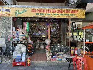 Đây là hình ảnh mặt tiền của một cửa hàng kinh doanh thiết bị điện tại địa chỉ 54 đường Hàng Đồng. Phía trên cửa hàng có một tấm bạt lớn màu vàng ghi dòng chữ "CHUYÊN BÁN THIẾT-BỊ ĐIỆN DÂN DỤNG - CÔNG NGHIỆP", "CÁP ĐIỆN 1 PHA - 3 PHA - ĐÈN CHIẾU SÁNG" và các số điện thoại liên hệ: ĐT: 02373.27 63.63, DĐ: 0942 57.63.63 - 0169.348.1207. Cửa hàng đang mở cửa, trưng bày rất nhiều sản phẩm cả bên trong và tràn ra vỉa hè. Các mặt hàng được bày bán rất đa dạng, bao gồm: nhiều loại đèn chiếu sáng (đèn LED dây, đèn trang trí, đèn đường, đèn pha năng lượng mặt trời), quạt điện, quạt thông gió công nghiệp, các cuộn dây cáp điện lớn, công tơ điện, và các thiết bị điện dân dụng khác. Phía trước cửa hàng có đặt một số bình chữa cháy và một hộp đựng dụng cụ PCCC màu đỏ có ghi số 114. Ngoài ra còn có một chiếc ghế nhựa màu đỏ và một chiếc bàn nhỏ.。（日本語訳）。これは、ハンドン通り54番地にある電設資材店の正面の写真です。店の上部には、「家庭用・産業用電設資材専門販売」、「単相・三相電力ケーブル - 照明器具」という文言と、連絡先電話番号：固定電話：02373.27 63.63、携帯電話：0942 57.63.63 - 0169.348.1207が書かれた大きな黄色のシートがあります。店は営業中で、店内にも歩道にもあふれるほど多くの商品が陳列されています。販売されている商品は非常に多様で、多くの種類の照明器具（LEDテープライト、装飾灯、街路灯、ソーラー投光器）、扇風機、産業用換気扇、大きな電線ケーブルの巻線、電力メーター、その他の家庭用電設資材が含まれます。店の前には、数本の消火器と、114と書かれた赤色の消防用具入れが置かれています。その他に、赤いプラスチックの椅子と小さなテーブルもあります。