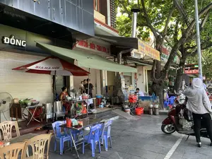 Bối cảnh là một quán ăn vỉa hè trên một con phố ở Thanh Hóa. Quán đang mở cửa phục vụ khách vào ban ngày. Nhiều người đang có mặt tại quán, bao gồm một gia đình (một người đàn ông, một người phụ nữ bế em bé) dường như đang đợi đồ ăn, một người phụ nữ mặc đồ hoa đang ngồi, và một người phụ nữ khác mặc áo chống nắng đứng cạnh xe máy. Hoạt động chính là buôn bán và ăn uống tại quán. Các vật thể nổi bật bao gồm bàn ghế nhựa và inox, một chiếc ô lớn có chữ "BIA HÀ NỘI", nhiều nồi inox lớn, bếp lò than với ống khói, và các xe máy đậu trên vỉa hè.。Các thông tin bằng chữ có thể nhận dạng được:。- Biển hiệu lớn trên tòa nhà: "ĐỒNG".。- Biển hiệu món ăn chữ đỏ: "BUN CHẢ - MIẾN - CHÁO LƯƠN, CÁ".。- Băng rôn quảng cáo màu vàng ghi địa chỉ "56 - HÀNG ĐỒNG", số điện thoại "ĐT: 0989.565.658", và các món ăn "MIẾN - CHÁO LƯƠN - BÚN CÁ - TRỨNG VỊT LỘN".。- Biển hiệu đèn LED ở phía sau: "THẮM HÙNG".。（日本語訳）。場面は、タインホアのある通りにある路上食堂です。店は日中に客を迎えて営業しています。店には多くの人がおり、その中には料理を待っていると思われる家族（男性一人、赤ちゃんを抱いた女性一人）、座っている花柄の服を着た女性、そして日焼け防止ジャケットを着てバイクの隣に立っている別の女性がいます。主な活動は、店での商売と飲食です。目立つ物には、プラスチックとステンレス製のテーブルと椅子、「BIA HÀ NỘI」という文字が入った大きな傘、多くの大きなステンレス製の鍋、煙突付きの炭火コンロ、そして歩道に停められたバイクが含まれます。識別できる文字情報：。- 建物にある大きな看板：「ĐỒNG」。- 赤い文字の料理の看板：「BUN CHẢ - MIẾN - CHÁO LƯƠN, CÁ」。- 住所「56 - HÀNG ĐỒNG」、電話番号「ĐT: 0989.565.658」、そして料理「MIẾN - CHÁO LƯƠN - BÚN CÁ - TRỨNG VỊT LỘN」が書かれた黄色の広告横断幕。- 後ろにあるLED看板：「THẮM HÙNG」。