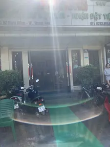 Bức ảnh chụp mặt tiền một nhà hàng vào ban ngày, bị lóa sáng mạnh.。**Chữ viết:**。- Trên biển hiệu chính của nhà hàng có các chữ, một phần bị lóa sáng: "... MÓN ĂN ĐẶC SẢN - NHẬN ĐẶT TIỆC".。- Địa chỉ trên biển hiệu: "Đ/C: 62 Hàng Đồng - TP. Thanh Hóa".。- Số điện thoại trên biển hiệu: "ĐT: 0373.851.6...".。- Biển số nhà gắn trên tường: "62 HÀNG ĐỒNG" và bên cạnh là "64 HÀNG ĐỒNG".。- Trên cột nhà số 62 có một tấm biển với khẩu hiệu: "NÓI LỜI HAY LÀM VIỆC TỐT HÀNH ĐỘNG THÂN THIỆN".。- Phía trên cửa ra vào có một dãy số điện thoại khác nhưng hơi mờ: "0978366314", "0868.464.345", ...。**Bối cảnh và hoạt động:**。- **Địa điểm:** Đây là lối vào của một nhà hàng nằm trên một con phố ở Thành phố Thanh Hóa.。- **Vật thể:** Phía trước cửa có một số xe máy và một chiếc xe đạp đang đỗ. Một chiếc ghế nhựa màu xanh lá cây được đặt ở góc trái ảnh. Lối vào có bậc tam cấp trải thảm xanh và cửa kính. Cây cảnh được trồng trong chậu đặt hai bên cửa.。- **Hoạt động:** Một người phụ nữ đang đứng ở bên phải lối vào. Nhìn qua cửa kính, có thể thấy một người khác đang ngồi bên trong nhà hàng, dường như đang sử dụng điện thoại. Khung cảnh cho thấy hoạt động thường ngày tại một cơ sở kinh doanh ăn uống.。（日本語訳）。日中に撮影されたレストランの正面の写真で、強い光で白飛びしている。**文字:**。- レストランのメイン看板には文字があり、一部が白飛びしている：「...名物料理 - 宴会予約承ります」。- 看板上の住所：「住所：タインホア市 ハンドン通り 62番地」。- 看板上の電話番号：「電話：0373.851.6...」。- 壁に取り付けられた番地プレート：「62 ハンドン」、その隣には「64 ハンドン」。- 62番地の家の柱には、「良い言葉を話し、良い行いをし、親切に行動する」というスローガンが書かれたプレートがある。- 出入り口の上には、別の電話番号の列があるが、少しぼやけている：「0978366314」、「0868.464.345」、...。**背景と活動:**。- **場所：** これはタインホア市のある通りに位置するレストランの入り口である。- **物体：** ドアの前には数台のバイクと一台の自転車が停まっている。入り口には緑のカーペットが敷かれた数段の階段とガラスのドアがある。- **活動：** 一人の女性が入り口の右側に立っている。ガラスのドア越しに見ると、レストランの中で別の人が座っており、どうやら電話を使っているようだ。この光景は、飲食店での日常の活動を示している。