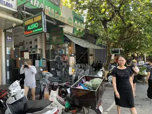 Bối cảnh là một con phố ở Việt Nam, cụ thể là trên đường Hàng Đồng. Có nhiều cửa hàng san sát nhau, trong đó nổi bật là một cửa hàng kinh doanh thiết bị điện tử và âm thanh.。Các dòng chữ có thể đọc được trên các biển hiệu bao gồm:。- Biển hiệu chính của cửa hàng: "HƯƠNG BINH AUDIO", địa chỉ "64 HÀNG ĐỒNG", và số điện thoại "ĐT: 0919 563 868".。- Biển số nhà màu xanh: "64 HÀNG ĐỒNG".。- Biển quảng cáo các sản phẩm: "ĐIỆN TỬ - ÂM THANH", "MÁY TRỢ GIẢNG", "ĐÀI RADIO + THẺ NHỚ", "MIC BLUETOOTH", "MIC LIVE STREAM".。- Cửa hàng bên cạnh có biển hiệu "ĐẠI LÝ ĐÈN LED, ĐỒ ĐIỆN VIỆT THU" tại "66 Hàng Đồng" và một biển nhỏ "ĐÓNG PIN, THIẾT BỊ CẦM TAY".。Hoạt động diễn ra trên phố khá nhộn nhịp. Một người phụ nữ mặc váy đen đang đi trên vỉa hè. Một người phụ nữ khác mặc đồ màu xám đang đứng gần cửa hàng và chỉnh lại khẩu trang. Ở phía sau, có một người phụ nữ đội nón lá đang bế một em bé.。Các vật thể đáng chú ý bao gồm:。- Nhiều loại loa thùng, quạt công nghiệp và các thiết bị điện tử khác được bày bán trước cửa hàng.。- Một số xe máy đang đậu trên vỉa hè. Trong đó có một chiếc xe máy được chế thêm một thùng gỗ lớn phía sau để chở hàng, bên trong thùng chứa đầy rau được bọc trong túi ni lông.。- Một cây xanh lớn có thân và cành được quấn dây đèn nháy trang trí.。（日本語訳）。背景はベトナムのある通り、具体的にはハンドン通りです。多くの店が隣接しており、その中で電子機器や音響機器を扱う店が目立っています。看板で読み取れる文字は以下の通りです：。- 店のメイン看板：「HƯƠNG BINH AUDIO」、住所「64 HÀNG ĐỒNG」、電話番号「ĐT: 0919 563 868」。- 青い番地プレート：「64 HÀNG ĐỒNG」。- 商品の広告看板：「電子機器 - 音響」、「講義補助マイク」、「ラジオ＋メモリーカード」、「Bluetoothマイク」、「ライブストリームマイク」。- 隣の店には「66 Hàng Đồng」にある「VIỆT THU LEDランプ・電気製品代理店」という看板と、「バッテリーパック、携帯機器」という小さな看板があります。通りでの活動はかなり賑やかです。黒いスカートを履いた女性が歩道を歩いています。別の灰色の服を着た女性が店の近くに立ち、マスクを直しています。後方には、ノンラーをかぶった女性が赤ちゃんを抱いています。注目すべき物体は以下の通りです：。- 店の前には、多くの種類のボックススピーカー、業務用扇風機、その他の電子機器が陳列販売されています。- 歩道には数台のバイクが停まっています。その中には、荷物を運ぶために後ろに大きな木箱が取り付けられたバイクがあり、箱の中はビニール袋に包まれた野菜でいっぱいです。- 大きな緑の木があり、その幹と枝には装飾用のイルミネーションライトが巻かれています。