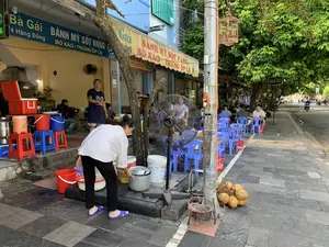 Bối cảnh là một quán ăn vỉa hè trên một con phố. Nhiều khách hàng đang ngồi ăn uống tại các bộ bàn ghế nhựa màu xanh được kê trên vỉa hè. Một chiếc quạt cây lớn đang hoạt động để làm mát cho khách. Một người phụ nữ đang cúi xuống làm việc bên cạnh một chiếc nồi nhôm lớn và nhiều xô nhựa. Bên trong quán, một người đàn ông mặc tạp dề đang đứng. Trên tường và mái hiên có các biển hiệu ghi tên quán là 'Bà Gái', địa chỉ '74 Hàng Đồng', và các món ăn như 'BÁNH MỲ SỐT VANG', 'BÒ XÀO - TRỨNG ỐP LA', và 'BIA HƠI'. Một biển hiệu khác treo trên cây ghi 'BIA 80'. Trên vỉa hè còn có một chùm dừa tươi, và một chiếc lồng chim được treo trên cột điện. Phía xa trên đường có xe cộ đang di chuyển.。（日本語訳）。舞台は、ある通りの路上飲食店です。多くの客が、歩道に置かれた青いプラスチック製のテーブルと椅子に座って飲食しています。客を涼しくするために、大きな扇風機が作動しています。一人の女性が、大きなアルミ鍋とたくさんのプラスチックのバケツの横で、かがんで作業をしています。店の中では、エプロンをつけた男性が立っています。壁と軒先には、店の名前「Bà Gái」、住所「74 Hàng Đồng」、そして「BÁNH MỲ SỐT VANG」、「BÒ XÀO - TRỨNG ỐP LA」、「BIA HƠI」といった料理名が書かれた看板があります。木に掛けられた別の看板には「BIA 80」と書かれています。歩道には、新鮮なココナッツの房もあり、電柱には鳥かごが吊るされています。遠くの道路では、車が行き交っています。