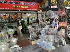 Bối cảnh là mặt tiền của một cửa hàng kinh doanh thiết bị điện. Trên biển hiệu chính màu đỏ có ghi 'CỬA HÀNG THIẾT BỊ ĐIỆN MINH HỒNG', địa chỉ tại '86 Hàng Đồng - Phường Điện Biên - TPTH' và số điện thoại '0918639592'. Cửa hàng trưng bày rất nhiều loại quạt điện (quạt cây, quạt treo tường, quạt bàn) để bán, nhiều chiếc còn mới nguyên trong bao bì ni lông. Hàng hóa được bày từ trong nhà ra đến vỉa hè. Có thể thấy các sản phẩm quạt của thương hiệu 'SENKO'. Một tấm biển nhỏ màu đỏ treo trên cây bên phải ghi 'BÁN ĐIỀU KHIỂN TIVI - QUẠT - ĐIỀU HÒA'. Thân cây này cũng được quấn quanh bằng dây đèn trang trí.。（日本語訳）。背景は電化製品店の正面です。赤いメイン看板には「MINH HONG電化製品店」、住所「86 Hàng Đồng - Điện Biên区 - TPTH」、電話番号「0918639592」と書かれています。店では、販売用に多くの種類の扇風機（スタンドファン、壁掛けファン、卓上ファン）が展示されており、多くはまだビニールのパッケージに入った新品です。商品は店内から歩道まで並べられています。「SENKO」ブランドの扇風機製品が見られます。右側の木に掛けられた小さな赤い看板には「テレビ・扇風機・エアコンのリモコン販売」と書かれています。この木の幹も装飾用のイルミネーションライトで巻かれています。