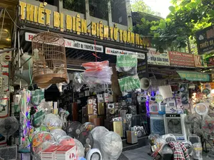 Đây là hình ảnh mặt tiền của một cửa hàng kinh doanh thiết bị điện và điện tử trên một con phố. Biển hiệu chính có chữ màu vàng nổi bật ghi 'THIẾT BỊ ĐIỆN - ĐIỆN TỬ TUẤN NGỌC'. Bên dưới là các biển hiệu nhỏ hơn với nội dung như 'ÂM THANH THIẾT BỊ TRUYỀN THANH', địa chỉ '88 HÀNG ĐỒNG', và 'ĐIỆN DÂN DỤNG'. Ngoài ra còn có biển hiệu của cửa hàng lân cận 'TOÀN TÙNG' tại số '90 HÀNG ĐỒNG', chuyên về 'K+, VTC-HD, THIẾT BỊ ÂM LY, LOA ĐÀI, TRUYỀN THANH'. Cửa hàng trưng bày vô số mặt hàng điện tử tràn ra cả lối đi, bao gồm rất nhiều loại quạt điện (quạt cây, quạt hộp), loa thùng, loa kéo di động, quạt điều hòa, đèn LED, đèn livestream, và các thiết bị âm thanh khác được đóng trong hộp hoặc bọc nilon. Phía trước cửa hàng có treo một chiếc lồng chim bằng gỗ. Một chiếc xe máy đậu phía trước, yên xe được phủ một tấm vải kẻ sọc.。（日本語訳）。これは、ある通りにある電気・電子機器を商う店の正面の写真です。メインの看板には「THIẾT BỊ ĐIỆN - ĐIỆN TỬ TUẤN NGỌC」と目立つ黄色の文字で書かれています。その下には、「音響・放送機器」、「88 HÀNG ĐỒNG」という住所、そして「家庭用電気製品」といった内容の小さな看板があります。また、「90 HÀNG ĐỒNG」番地にある隣の店「TOÀN TÙNG」の看板もあり、「K+、VTC-HD、アンプ機器、スピーカー、放送」を専門としています。店は通路にまで溢れ出るほどの無数の電子製品を陳列しており、それには多くの種類の扇風機（スタンドファン、ボックスファン）、スピーカー、ポータブルスピーカー、冷風扇、LEDライト、ライブ配信用ライト、そして箱詰めまたはビニールで包まれたその他の音響機器が含まれています。店の前には木製の鳥かごが一つ吊るされています。前方にはバイクが一台停まっており、そのシートは縞模様の布で覆われています。