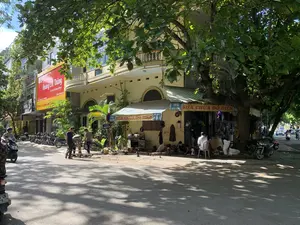 Bức ảnh chụp một góc phố tại thành phố Thanh Hóa vào ban ngày, dưới bóng râm của một cây lớn. Dọc theo con đường là các tòa nhà, trong đó có một tòa nhà với tấm biển quảng cáo lớn màu đỏ ghi 'Hoàng Cốt Thông', 'Trị liệu Cơ Xương Khớp', địa chỉ '51 Nguyễn Du - TP Thanh Hóa' và hotline '0866 205 665'. Tại góc đường là một cửa hàng sửa chữa đồ điện với biển hiệu '92 HÀNG ĐỒNG SỬA CHỮA ĐỒ ĐIỆN'. Một biển báo đường phố cũng xác nhận tên đường là 'ĐƯỜNG NGUYỄN DU'. Trên vỉa hè, trước cửa hàng sửa chữa, có một vài người đàn ông đang làm việc; một người ngồi xổm sửa chữa thiết bị, một người khác ngồi trên ghế nhựa. Một nhóm người khác đang đứng cạnh một chiếc xe đẩy. Nhiều xe máy đang được đậu trên vỉa hè và có người đang tham gia giao thông trên đường.。（日本語訳）。写真は、日中、大きな木の木陰にあるタインホア市のある街角を写したものです。道沿いには建物が並んでおり、その中の一つの建物には、「Hoàng Cốt Thông」、「Trị liệu Cơ Xương Khớp」、住所「51 Nguyễn Du - TP Thanh Hóa」、ホットライン「0866 205 665」と書かれた大きな赤い広告看板があります。道の角には、「92 HÀNG ĐỒNG SỬA CHỮA ĐỒ ĐIỆN」という看板のある電化製品の修理店があります。ある道路標識も、通りの名前が「ĐƯỜNG NGUYỄN DU」であることを示しています。歩道の上、修理店の前では、数人の男性が作業をしています。一人はしゃがんで機器を修理しており、もう一人はプラスチックの椅子に座っています。別のグループの人々が手押し車の隣に立っています。多くのバイクが歩道に停められており、道路では人々が通行しています。