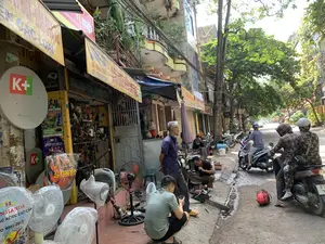 Đây là khung cảnh trên một con phố ở đô thị Việt Nam, có thể là thành phố Thanh Hóa, với các cửa hàng và nhà ở san sát nhau. Bên trái là một cửa hàng sửa chữa và buôn bán đồ điện, với nhiều loại quạt điện (quạt cây, quạt hộp) được bày bán trên vỉa hè. Một số quạt có thương hiệu 'HANJIANDA' và 'SENKO'. Trên vỉa hè, một nhóm đàn ông đang tụ tập. Một người đang ngồi sửa một chiếc máy cưa, một người khác ngồi xổm xem điện thoại, và một người đàn ông lớn tuổi đang đứng quan sát. Trên đường có nhiều xe máy đang di chuyển, một số người mặc áo chống nắng và đội mũ bảo hiểm. Các biển hiệu có thể đọc được bao gồm: biển hiệu cửa hàng màu vàng ghi 'SỬA CHỮA ĐIỆN CƠ... CÁC LOẠI' và số điện thoại 'ĐT: 0384.541.578'; các chảo vệ tinh và áp phích quảng cáo của truyền hình K+ cho giải đấu 'UEFA EURO 2024'; một biển chỉ dẫn màu đỏ ghi 'Xương Khớp' 'CÁCH 50M'; và trên hộp quạt có các dòng chữ 'QUẠT SẠC AC/DC CAO CẤP' và 'ISO 9001:2015'. Phía trên là hệ thống dây điện chằng chịt và cây xanh ven đường.。（日本語訳）。これはベトナムの都市、おそらくタインホア市の通りでの光景で、店や家が密集しています。左側には電化製品の修理・販売店があり、多くの種類の扇風機（スタンドファン、ボックスファン）が歩道に陳列されています。いくつかの扇風機は「HANJIANDA」や「SENKO」というブランドのものです。歩道では、男性のグループが集まっています。一人は座ってチェーンソーを修理しており、別の人はしゃがんで携帯電話を見ており、そして年配の男性が立って見守っています。路上では多くのバイクが走行しており、何人かの人々は日焼け防止の上着を着てヘルメットをかぶっています。読み取れる看板には、「SỬA CHỮA ĐIỆN CƠ... CÁC LOẠI」（電動機...各種修理）と電話番号「ĐT: 0384.541.578」が書かれた黄色い店の看板、「UEFA EURO 2024」大会のためのK+テレビの衛星放送パラボラアンテナと広告ポスター、「Xương Khớp」（骨関節）「CÁCH 50M」（50m先）と書かれた赤い案内標識、そして扇風機の箱にある「QUẠT SẠC AC/DC CAO CẤP」（高級AC/DC充電式扇風機）と「ISO 9001:2015」という文字が含まれます。上方には、複雑に絡み合った電線網と街路樹があります。