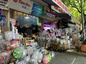 Bối cảnh là một cửa hàng bán đồ điện tử và điện dân dụng trên một con phố tại thành phố Thanh Hóa. Rất nhiều quạt điện các loại (quạt cây, quạt bàn), vẫn còn mới và được bọc trong túi ni lông, được bày bán trên vỉa hè trước cửa hàng. Bên trong cửa hàng, có một người đang ngồi, và cửa hàng được trang trí bằng dây đèn nháy nhiều màu và một chiếc đèn lồng đỏ.。Các thông tin văn bản trên các biển hiệu được ghi lại như sau:。- Biển hiệu chính của cửa hàng: "ĐẠI LÝ HÀ SINH - ĐIỆN TỬ, ĐIỆN DÂN DỤNG, THIẾT BỊ PHÁT THANH TRUYỀN HÌNH. Chuyên cung cấp các sản phẩm Nguồn sáng & Thiết bị chiếu sáng. Đ/C: Số nhà 96 Hàng Đồng, P. Điện Biên, Tp. Thanh Hóa. ĐT: 02373.753.737 - 0916.797.314".。- Phía trên có biển hiệu của thương hiệu "Rạng Đông" với logo và mã QR.。- Một biển hiệu treo ngang màu đỏ và trắng có chữ "SỬA CHỮA ĐỒ ĐIỆN", "THẾ GIỚI ĐIỆN" và "96 HÀNG ĐỒNG".。- Biển hiệu của cửa hàng lân cận: "CHIẾN HÒA - NNP THIẾT BỊ ĐIỆN: LIOA - STANDA... HÀNG ĐỒNG - P. ĐIỆN BIÊN - TP THANH HÓA".。- Các biển nhỏ khác có nội dung: "BÁN ĐIỀU KHIỂN CÁC LOẠI" và "NHẬN BÁN VÀ SỬA CHỮA ... KHIỂN".。Hoạt động chính là kinh doanh, buôn bán các thiết bị điện. Các vật thể khác có thể thấy bao gồm loa phóng thanh, các hộp sản phẩm, một cây xanh và một cột điện trên vỉa hè.。（日本語訳）。背景は、タインホア市のある通りにある電化製品と家庭用電化製品の店です。多くの種類の扇風機（スタンドファン、卓上ファン）が、まだ新品でビニール袋に包まれたまま、店の前の歩道に陳列販売されています。店内には一人の人が座っており、店は色とりどりのイルミネーションライトと赤い提灯で飾られています。看板に書かれているテキスト情報は以下の通りです。- 店のメイン看板：「ハーシン代理店 - 電子機器、家庭用電化製品、放送・テレビ設備。光源製品＆照明器具の専門供給。住所：タインホア市ディエンビエン区ハンドン通り96番地。電話：02373.753.737 - 0916.797.314」。- 上部には、ロゴとQRコードが付いた「Rạng Đông（ラン・ドン）」ブランドの看板があります。- 「電化製品修理」、「電気の世界」、「ハンドン96」という文字が入った赤と白の横長の看板があります。- 隣の店の看板：「CHIẾN HÒA（チエン・ホア） - NNP電気設備：LIOA - STANDA... ハンドン - ディエンビエン区 - タインホア市」。- その他の小さな看板には、「各種リモコン販売」および「販売および修理受付...コン」という内容が書かれています。主な活動は、電気設備の商売、販売です。その他に見られる物には、拡声器、製品の箱、緑の木、歩道上の電柱などがあります。