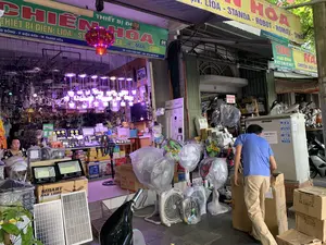 Đây là khung cảnh phía trước một cửa hàng thiết bị điện trên đường phố ở thành phố Thanh Hóa. Cửa hàng trưng bày rất nhiều mặt hàng, từ bên trong ra đến vỉa hè. Các biển hiệu cho thấy tên cửa hàng có thể là "CHIẾN HÒA", chuyên kinh doanh thiết bị điện, điện cơ của các hãng như LIOA, STANDA, ROBOT, ROMAN, SINO. Địa chỉ được ghi trên biển là số 98 và 100 đường Hàng Đồng, Phường Điện Biên, TP. Thanh Hóa, cùng với các số điện thoại liên lạc như 02373 852 989 và 0912 00 99 89. Các sản phẩm được bày bán rất đa dạng, bao gồm nhiều loại đèn (đèn LED, đèn pha, đèn trang trí), quạt điện (quạt cây, quạt hộp), tấm pin năng lượng mặt trời, và các thiết bị điện khác, nhiều sản phẩm vẫn còn trong bao bì. Có một người phụ nữ đang ngồi bên trong quầy hàng và một người đàn ông mặc áo xanh đang sắp xếp hàng hóa trong các thùng carton ở bên ngoài. Một chiếc đèn lồng đỏ được treo trang trí. Phía trước có một chiếc xe máy đang đỗ. Trên một thiết bị lớn màu trắng có dán giấy cảnh báo "CẢM TRÈO。ĐIỆN ÁP CAO NGUY HIỂM" và các sticker quảng cáo dịch vụ "HÚT BỂ PHỐT".。（日本語訳）。これはタインホア市の路上にある電化製品店の前の光景です。店は店内から歩道まで、たくさんの商品を陳列しています。看板によると、店名は「CHIẾN HÒA（チエン・ホア）」のようで、LIOA、STANDA、ROBOT、ROMAN、SINOなどのメーカーの電気設備、電動機を専門に扱っています。看板に記載されている住所はタインホア市ディエンビエン区ハンドン通り98番と100番で、連絡先の電話番号は02373 852 989と0912 00 99 89です。販売されている商品は非常に多様で、多くの種類の照明（LEDライト、投光器、装飾灯）、扇風機（スタンドファン、ボックスファン）、太陽光パネル、その他の電気機器が含まれており、多くの商品はまだ梱包されたままです。店内のカウンターには女性が一人座っており、外では青いシャツを着た男性が段ボール箱の中の商品を整理しています。赤い提灯が一つ、装飾として吊るされています。前方にはバイクが一台停まっています。大きな白い機器には「登るな。高電圧危険」という警告紙と、「浄化槽清掃」サービスの広告ステッカーが貼られています。