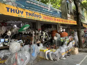 Bức ảnh chụp mặt tiền của một cửa hàng kinh doanh thiết bị điện trên một con phố. Cửa hàng có tên là 'THỦY THẮNG', địa chỉ tại '102 - HÀNG ĐỒNG'. Các biển hiệu cho thấy cửa hàng chuyên về 'ĐIỆN CÔNG NGHIỆP - ĐIỆN DÂN DỤNG - AUTOMAT - KHỞI ĐỘNG TỪ' và 'CÁP ĐIỆN ĐỒNG - NHÔM'. Một biển hiệu khác ở trên quảng cáo 'DÂY VÀ CÁP ĐIỆN GOLDCUP'. Rất nhiều hàng hóa được trưng bày bên trong và tràn ra cả vỉa hè, bao gồm nhiều loại quạt điện mới (quạt cây, quạt treo tường, quạt hộp) của các thương hiệu như SENKO, vẫn còn được bọc trong túi ni lông. Ngoài ra còn có các loại đèn chiếu sáng (đèn đường, đèn pha LED), các cuộn lớn ống luồn dây điện màu cam, và các thiết bị điện công nghiệp khác. Có một cái cây lớn ở bên phải vỉa hè. Hoạt động chính là buôn bán, với một vài người có mặt tại cửa hàng.。（日本語訳）。ある通りにある電気設備を販売する店の正面を撮影した写真です。店の名前は「THỦY THẮNG」、住所は「102 - HÀNG ĐỒNG」です。看板には、この店が「産業用電気 - 民生用電気 - サーキットブレーカー - 電磁開閉器」および「銅・アルミニウム電線」を専門としていることが示されています。上にある別の看板は「GOLDCUP電線・ケーブル」を宣伝しています。多くの商品が店内や歩道にまであふれるように陳列されており、その中にはSENKOなどのブランドの新品の扇風機（スタンドファン、壁掛けファン、ボックスファン）が多数含まれ、まだビニール袋に包まれています。その他にも、各種照明器具（街路灯、LED投光器）、オレンジ色の電線管の大きな巻物、その他の産業用電気設備があります。歩道の右側には大きな木が一本あります。主な活動は販売で、店には数人の人がいます。