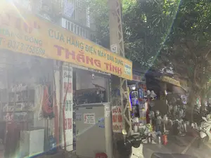 Bối cảnh là một con phố với các cửa hàng kinh doanh san sát nhau, có thể là một khu chợ hoặc dãy phố thương mại. Nổi bật là một cửa hàng bán đồ điện máy dân dụng có tên "THẮNG TÍNH" tại địa chỉ 116b.。Các thông tin văn bản có thể đọc được bao gồm:。- Trên biển hiệu chính màu vàng: "CỬA HÀNG ĐIỆN MÁY DÂN DỤNG THẮNG TÍNH", địa chỉ "116b", và các số điện thoại "ĐT: 0373.758680", "ĐT: 0936 434 000".。- Trên tường và cột điện có các biển quảng cáo nhỏ hơn: "SỬA MÁY HÀN ĐIỆN TỬ", "DƯƠNG HOÀNG Chuyên PHỤ KIỆN ĐIỆN THOẠI".。- Trên một tủ điện màu xám có dán cảnh báo nguy hiểm: "CẤM LẠI GẦN。CÓ ĐIỆN NGUY HIỂM CHẾT NGƯỜI".。- Nhiều mẩu quảng cáo dán trên cột điện và tủ điện cho dịch vụ "HÚT BỂ PHỐT" với các số điện thoại như 0989.171.353, 0942.85.23.85, 0941.597.345, 0974.61.42.52.。- Cửa hàng bên cạnh có tên "ĐIỆN TÙNG LIÊN", bán các sản phẩm của "LIOA - STANOA - SINO".。Hoạt động chính là kinh doanh, các cửa hàng đang mở cửa và trưng bày hàng hóa ra vỉa hè để bán. Các vật thể có trong ảnh bao gồm: rất nhiều quạt điện các loại (quạt cây, quạt treo tường) được bày bán phía trước, các cuộn dây điện, kệ hàng chứa sản phẩm bên trong cửa hàng, một cột điện bê tông lớn, một tủ điện, một chiếc xe máy tay ga màu trắng đang đỗ, và cây xanh có tán lá rậm rạp.。（日本語訳）。背景は、商店が隣接して並んでいる通りで、市場か商店街のようです。目立っているのは、住所116bにある「THẮNG TÍNH」という名前の家庭用電化製品店です。読み取れる文字情報には、以下のものが含まれます：。- 黄色いメインの看板：「家庭用電化製品店 THẮNG TÍNH」、住所「116b」、および電話番号「電話：0373.758680」、「電話：0936 434 000」。- 壁や電柱にある小さな広告看板：「電子溶接機修理」、「DƯƠNG HOÀNG 携帯電話アクセサリー専門」。- 灰色の配電盤に貼られた危険警告：「近づくな。感電死の危険あり」。- 電柱や配電盤に貼られた「浄化槽吸引」サービスの多数の広告には、0989.171.353、0942.85.23.85、0941.597.345、0974.61.42.52などの電話番号が記載されています。- 隣の店は「ĐIỆN TÙNG LIÊN」という名前で、「LIOA - STANOA - SINO」の製品を販売しています。主な活動は商売で、各店は開店しており、商品を歩道に陳列して販売しています。写真に写っている物には、前面に陳列販売されている多くの種類の扇風機（スタンド扇風機、壁掛け扇風機）、電線の巻線、店内の商品を収めた棚、大きなコンクリートの電柱、配電盤、駐車している白いスクーター、そして葉が茂った緑の木が含まれます。