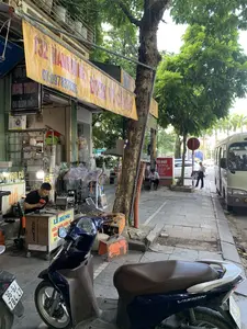 Bức ảnh chụp cảnh sinh hoạt trên vỉa hè của một con phố ở đô thị. Ở phía trước bên trái, một người đàn ông mặc áo đen đang ngồi tại một quầy sửa chữa đồ điện tử tạm bợ. Trên quầy có biển hiệu 'LÊ HÙNG SỬA CHỮA ĐIỆN TỬ'. Xung quanh anh ta là các thiết bị như máy hàn (có chữ 'ARC200D', 'LGwelder'), quạt điện, vợt muỗi điện và các linh kiện khác. Phía trên cửa hàng có một tấm bạt quảng cáo lớn màu vàng đã cũ, ghi địa chỉ '132 MẠNH DŨNG', số điện thoại 'ĐT:0978378385' và dòng chữ 'SỬA CHỮA...'. Bên cạnh có biển hiệu 'HÙNG THANH 134 HÀNG ĐỒNG' và biển số nhà '134'. Ở phía sau, dưới bóng cây, có một quán cà phê với biển hiệu đỏ 'CÀ PHÊ' và thực đơn đồ uống. Hai người đàn ông đang ngồi trên ghế ở quán. Trên vỉa hè có đỗ vài chiếc xe máy, trong đó có một chiếc Honda Vision màu xanh tím. Một chiếc xe buýt nhỏ màu trắng đang đỗ bên lề đường. Một người phụ nữ đang đi bộ trên vỉa hè và có một biển báo giao thông hình tròn màu đỏ cấm dừng đỗ.。（日本語訳）。都市のある通りの歩道での生活風景を撮った写真です。左手前には、黒い服を着た男性が、間に合わせの電子機器修理店に座っています。店先には「LÊ HÙNG SỬA CHỮA ĐIỆN TỬ」という看板があります。彼の周りには、溶接機（「ARC200D」「LGwelder」の文字あり）、扇風機、電撃殺虫ラケット、その他の部品などの機器があります。店の上には、古びた黄色の大きな広告シートがあり、「132 MẠNH DŨNG」という住所、「ĐT:0978378385」という電話番号、そして「SỬA CHỮA...」という文字が書かれています。隣には「HÙNG THANH 134 HÀNG ĐỒNG」という看板と、「134」という番地のプレートがあります。後方の木陰には、「CÀ PHÊ」と書かれた赤い看板とドリンクメニューがあるカフェがあります。二人の男性がその店で椅子に座っています。歩道には数台のバイクが停まっており、その中には青紫色のホンダ・ビジョンが1台あります。白い小型バスが1台、道端に停まっています。一人の女性が歩道を歩いており、駐停車を禁止する赤い円形の交通標識があります。