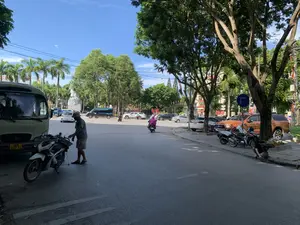 Quang cảnh một ngã tư hoặc bùng binh tại một thành phố ở Việt Nam, có khả năng là thành phố Thanh Hóa, vào một ngày nắng đẹp. Bầu trời trong xanh, hai bên đường trồng nhiều cây xanh lớn và cây cọ.。Ở trung tâm của bùng binh, có một tượng đài lớn bằng đá. Nhiều phương tiện giao thông như ô tô và xe máy đang lưu thông trên đường. Một người phụ nữ mặc áo hồng đang điều khiển xe máy.。Ở phía trước bên trái, một người đàn ông đội mũ, mặc áo phông và quần short đang đứng cạnh một chiếc xe máy. Gần đó là một chiếc xe tải nhỏ màu trắng có ghi dòng chữ 'XE HỢP ĐỒNG'.。Ở vỉa hè bên phải, một số xe máy đang đậu và có một người đàn ông đang ngồi xổm làm việc gần một gốc cây. Có một biển báo giao thông hình tròn màu xanh, chỉ dẫn hướng đi bắt buộc rẽ phải. Phía xa có các tòa nhà và những lá cờ đỏ được treo.。（日本語訳）。ベトナムのある都市、おそらくはタインホア市の、ある晴れた日の交差点またはロータリーの光景。空は澄み切って青く、道の両側には大きな緑の木々とヤシの木がたくさん植えられている。ロータリーの中央には、石でできた大きな記念碑がある。自動車やバイクなど多くの交通車両が道路を通行している。ピンクの服を着た一人の女性がバイクを運転している。左手前には、帽子をかぶり、Tシャツとショートパンツを履いた男性がバイクの隣に立っている。その近くには、「XE HỢP ĐỒNG」という文字が書かれた白い小型トラックがある。右側の歩道には、数台のバイクが駐車しており、一人の男性が木の根元近くでしゃがんで作業をしている。右折必須の進行方向を示す、青い円形の交通標識がある。