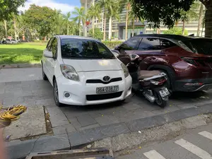 Bức ảnh chụp cảnh các phương tiện đang đỗ trên vỉa hè lát gạch trước một tòa nhà lớn. Ở phía sau, trên tòa nhà có một tấm biển lớn ghi chữ: "TRUNG TÂM PHỤC VỤ HÀNH CHÍNH CÔNG TỈNH THANH HÓA" và "SỐ 28 ĐẠI LỘ LÊ LỢI - PHƯỜNG ĐIỆN BIÊN - TP. THANH HÓA". Ở cận cảnh, một chiếc ô tô màu trắng hiệu Toyota đang đỗ. Bên cạnh nó là một chiếc xe máy tay ga màu nâu và một chiếc ô tô SUV màu đỏ mận. Xung quanh là cây xanh, bao gồm cả những cây cọ cao, và một bãi cỏ. Một lá cờ Việt Nam được treo trên tòa nhà. Dưới đất có một cuộn dây điện màu vàng.。（日本語訳）。大きな建物の前にあるタイル敷きの歩道に車両が駐車している様子を写した写真です。後方、建物には「TRUNG TÂM PHỤC VỤ HÀNH CHÍNH CÔNG TỈNH THANH HÓA」と「SỐ 28 ĐẠI LỘ LÊ LỢI - PHƯỜNG ĐIỆN BIÊN - TP. THANH HÓA」と書かれた大きな看板があります。手前には、トヨタ製の白い自動車が駐車しています。その隣には、茶色のスクーターとワインレッドのSUV車があります。周囲には、高いヤシの木を含む緑の木々と芝生があります。建物にはベトナムの国旗が掲げられています。地面には黄色い電線のコイルが一つあります。