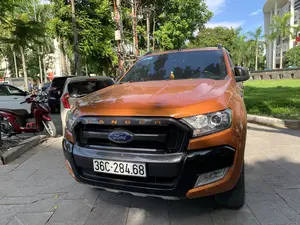 Hình ảnh chụp một chiếc xe bán tải hiệu Ford Ranger màu cam đang đỗ trên vỉa hè lát đá. Trên lưới tản nhiệt phía trước của xe có dòng chữ 'R A N G E R' và logo 'Ford'. Bối cảnh là một không gian đô thị, có thể là ven một con đường hoặc trong một bãi đỗ xe ở thành phố. Xung quanh có nhiều cây xanh tươi tốt, bao gồm cả cây cọ, và các cột điện với hệ thống dây điện chằng chịt. Ở phía sau và bên cạnh, có thể thấy các phương tiện khác như ô tô và xe máy cũng đang đỗ. Phía xa bên phải là một bãi cỏ và một phần của một tòa nhà lớn, có kiến trúc hiện đại. Hoạt động chính trong ảnh là các phương tiện đang đỗ.。（日本語訳）。石畳の歩道に駐車しているオレンジ色のフォード・レンジャーのピックアップトラックを撮影した写真。車のフロントグリルには「R A N G E R」の文字と「Ford」のロゴがある。背景は都市空間で、道路脇か市内の駐車場かもしれない。周囲にはヤシの木を含む青々とした緑の木々が多く、電線が複雑に絡み合った電柱がある。写真の中での主な活動は、車両が駐車していることである。