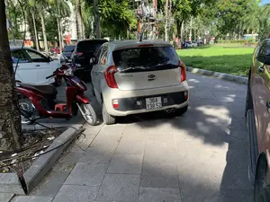 Cảnh quan đô thị chụp tại một vỉa hè lát đá. Nhiều phương tiện đang đỗ trên vỉa hè, bao gồm một chiếc ô tô màu be hiệu KIA Morning ở vị trí trung tâm. Có thể thấy chữ "KIA" và "morning" trên xe. Bên cạnh chiếc ô tô này là một chiếc xe máy tay ga màu đỏ, có thể nhận ra chữ "VISION". Xung quanh còn có các ô tô khác đang đỗ. Bên phải là một khu vực trồng cỏ và cây xanh, có thể là công viên hoặc dải phân cách. Phía sau là hàng cây xanh, bao gồm cả cây cọ, và một con đường có phương tiện đang lưu thông. Có một cột điện với trạm biến áp và hệ thống dây điện. Ở phía xa có một biểu ngữ màu đỏ nhưng không thể đọc rõ chữ.。（日本語訳）。石畳の歩道で撮影された都市景観。多くの車両が歩道に駐車しており、中央にはKIAモーニングというブランドのベージュ色の自動車が1台含まれている。この自動車の隣には赤いスクーターが1台あり、「VISION」の文字が認識できる。周りには他の自動車も駐車している。右側には草木が植えられたエリアがあり、公園または中央分離帯の可能性がある。後方にはヤシの木を含む並木と、車両が通行している道路がある。変圧器と配電システムを備えた電柱が1本ある。遠方には赤い横断幕があるが、文字ははっきりと読み取れない。