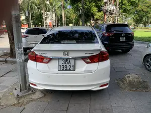 Bức ảnh chụp lại một khung cảnh trên vỉa hè của một con đường đô thị, có nhiều cây xanh bao gồm cả cây cọ. Hoạt động chính là những chiếc ô tô đang đỗ. Ở vị trí trung tâm là phía sau của một chiếc ô tô Honda City màu trắng. Trên xe có các dòng chữ 'CITY' và 'i-VTEC'. Phía sau bên phải có dán một tem nhỏ của 'Garage ôtô Hải Liên' với các thông tin như 'PHÒNG SƠN HẤP' và 'SƠN VI TÍNH'. Đỗ gần đó là một chiếc Mazda CX-5 màu đen và một chiếc Mazda 2 màu trắng. Có thể đọc được chữ 'CX-5' trên xe màu đen và 'SPORT' trên xe Mazda 2. Bối cảnh phía sau là đường phố, cây cối, một bãi cỏ và một phần tòa nhà. Có thể thấy một vài lá cờ đỏ ở phía xa. Ngoài ra còn có các cột điện, một trạm biến áp và một con thằn lằn nhỏ ở chân cột kim loại.。（日本語訳）。写真は、ヤシの木を含む多くの緑樹がある都市の道路の歩道上の光景を捉えたものです。主な活動は駐車している車です。中央には、白いホンダ・シティの後部があります。車には「CITY」と「i-VTEC」の文字があります。後部右側には、「PHÒNG SƠN HẤP」や「SƠN VI TÍNH」といった情報が記載された「Garage ôtô Hải Liên」の小さなステッカーが貼られています。その近くには黒いマツダ・CX-5と白いマツダ・2が駐車しています。黒い車からは「CX-5」、マツダ・2からは「SPORT」の文字が読み取れます。背景は、道路、木々、芝生、そして建物の一部です。遠方には、いくつかの赤い旗が見えます。その他にも、電柱、変電設備、そして金属製の柱の根元にいる小さなトカゲがいます。