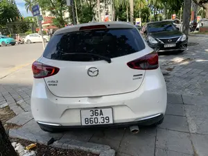 Bức ảnh chụp một chiếc ô tô màu trắng hiệu Mazda 2 Sport, phiên bản Luxury với công nghệ Skyactiv-G, đang đỗ trên vỉa hè của một con phố. Vỉa hè được lát bằng gạch đá. Xung quanh có nhiều cây xanh, bao gồm cả cây cọ. Ở phía trước, một chiếc ô tô màu đen khác cũng đang đỗ. Trên đường có các phương tiện khác đang di chuyển, trong đó có một chiếc xe máy có hai người ngồi. Một biển báo giao thông màu xanh lam có thể được nhìn thấy với dòng chữ "ĐƯỜNG HÀNG ĐỒNG". Bối cảnh cho thấy đây là một khu vực đô thị vào ban ngày.。（日本語訳）。歩道は石のタイルで舗装されています。周りにはヤシの木を含む多くの緑の木があります。前方には、別の黒い車も駐車しています。道路上では他の車両が移動しており、その中には二人乗りのバイクが一台います。「ĐƯỜNG HÀNG ĐỒNG」と書かれた青い交通標識が見えます。背景から、ここが日中の都市部であることがわかります。