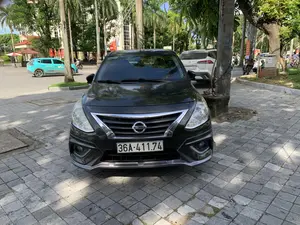 Bức ảnh chụp một chiếc ô tô màu đen hiệu Nissan đang đỗ trên vỉa hè lát đá của một con đường trong thành phố. Vỉa hè có nhiều cây xanh, bao gồm cả cây cọ. Phía sau chiếc xe Nissan là một chiếc ô tô màu trắng hiệu Toyota Corolla Cross. Trên đường phố, có các phương tiện giao thông khác đang hoạt động, bao gồm một chiếc taxi điện VinFast màu xanh lam của hãng Xanh SM và một vài người đang đi xe máy. Trên thân chiếc taxi Xanh SM có thể đọc được dòng chữ 'XANH SM' và số điện thoại '1900 2088'. Ở phía xa, có các tòa nhà, một cổng vào với chốt bảo vệ mái ngói đỏ và nhiều lá cờ đỏ được treo. Có một cột điện với các số '091 525 119' và một tủ điện ghi 'TC1.1'. Một biển báo giao thông cấm đi ngược chiều cũng xuất hiện trong khung cảnh.。（日本語訳）。歩道にはヤシの木を含む多くの緑の木があります。日産車の後ろには、トヨタ・カローラクロスブランドの白い車があります。路上では、Xanh SM社の青いVinFast製電気タクシーや、バイクに乗っている数人の人々など、他の交通車両が走行しています。Xanh SMタクシーの車体には、「XANH SM」という文字と電話番号「1900 2088」が読み取れます。遠くには、建物、赤い瓦屋根の警備所がある入口の門、そして掲げられた多くの赤い旗があります。「091 525 119」という数字が書かれた電柱と、「TC1.1」と記された配電盤があります。逆走を禁止する交通標識も風景の中に現れています。