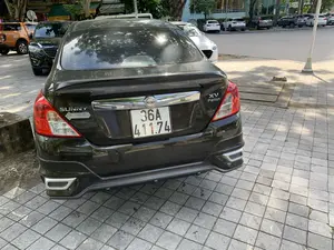 Bức ảnh chụp phía sau của một chiếc ô tô màu đen hiệu Nissan Sunny, phiên bản XV Premium, đang đỗ trên vỉa hè lát gạch. Trên thân xe có các dòng chữ 'SUNNY', 'XV Premium', logo 'NISSAN' và một nhãn dán ghi 'THĂNG LONG'. Bối cảnh là một con đường đô thị, có nhiều cây xanh. Ở phía sau và bên kia đường, có nhiều ô tô khác đang đỗ. Có thể nhìn thấy các biển hiệu của cửa hàng và tòa nhà, bao gồm 'NG DỊCH VỤ CẦM ĐỒ', một phần biển hiệu 'THỰC PHẨM HỮU CƠ' và logo ngân hàng 'BIDV'.。（日本語訳）。タイル張りの歩道に駐車している、日産サニーXVプレミアムバージョンの黒い自動車の後ろ姿の写真。車体には「SUNNY」、「XV Premium」の文字、「NISSAN」のロゴ、そして「THĂNG LONG」と書かれたステッカーがある。背景は緑の多い都市の道路である。後方と道路の向かい側には、他の多くの車が駐車している。