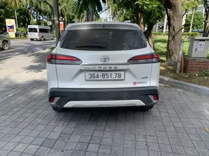 Bức ảnh chụp phía sau của một chiếc ô tô SUV màu trắng, hiệu Toyota Corolla Cross, đang đỗ trên vỉa hè lát gạch đá màu xám. Kính sau và thân xe có bám bụi. Trên đuôi xe có các dòng chữ "COROLLA CROSS", "1.8V" và logo Toyota. Có một nhãn dán của đại lý "TOYOTA THANH HÓA" với thông tin "Số 253 Trần Phú - P. Ba Đình - TP. Thanh Hóa" và trang web. Bên phải chiếc xe là một tủ điện màu xám, trên đó có ghi "TC1.1", "TBA NGỌC TRẠO", và "TỔNG ĐÀI CSKH 1900 8799". Khung cảnh xung quanh là một con đường đô thị với cây xanh, bao gồm cả cây cọ. Ở phía xa, có một chiếc xe van màu bạc đang di chuyển trên đường và một biển quảng cáo nhưng không thể đọc rõ nội dung.。（日本語訳）。灰色の石タイルで舗装された歩道に駐車している、トヨタ・カローラクロスというブランドの白いSUV車の後方を撮影した写真。リアガラスと車体には埃が付着している。車のリア部分には「COROLLA CROSS」、「1.8V」という文字とトヨタのロゴがある。「TOYOTA THANH HÓA」というディーラーのステッカーがあり、「タインホア市バーディン区チャンフー通り253番地」という情報とウェブサイトが記載されている。車の右側には灰色の配電盤があり、そこには「TC1.1」、「TBA NGỌC TRẠO」、そして「カスタマーケアコールセンター 1900 8799」と書かれている。周囲の風景は都市の道路で、ヤシの木を含む緑の木々がある。遠くには、銀色のバンが1台道路を走行しており、広告看板があるが、その内容ははっきりと読み取れない。