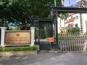 Bức ảnh chụp cổng chính của một cơ quan công quyền, đó là Trụ sở Ủy ban Nhân dân tỉnh Thanh Hóa. Khung cảnh cho thấy một ngày nắng đẹp.。Các thông tin bằng chữ viết có thể nhận dạng được:。1. Biển hiệu chính có nền vàng, chữ đỏ ghi: "TRỤ SỞ ỦY BAN NHÂN DÂN TỈNH THANH HÓA". Phía dưới là thông tin địa chỉ, điện thoại và website: "ĐC: SỐ 35 ĐẠI LỘ LÊ LỢI, P. LAM SƠN, TP. THANH HÓA ĐT: 0237 3852 246 - Website://www.thanhhoa.gov.vn".。2. Một biển báo nhỏ màu đỏ gắn trên cổng ghi: "CẤM GHI ÂM, GHI HÌNH CHỤP ẢNH" và bản dịch tiếng Anh "NO RECORDING, FILMING, PHOTO - TAKING".。3. Trên tòa nhà ở phía sau có một băng rôn màu đỏ với chữ vàng, một phần có thể đọc được là "...NƯỚC CỘNG HÒA XÃ HỘI CHỦ NGHĨA VIỆT NAM MUÔN NĂM".。Miêu tả cảnh vật và vật thể:。- Địa điểm là một tòa nhà văn phòng chính phủ.。- Có một cổng sắt lớn màu đen với các chi tiết trang trí và một hàng rào sắt bao quanh. Bên cạnh đó là một cổng xếp bằng inox có thể co giãn.。- Phía sau cổng là một tòa nhà lớn, màu trắng, nhiều tầng. Trên mặt tiền của tòa nhà có gắn Quốc huy Việt Nam.。- Có nhiều cây xanh lớn và các bụi cây nhỏ được cắt tỉa gọn gàng.。- Nhiều lá cờ, bao gồm cờ Việt Nam và cờ Đảng, đang bay trên đỉnh cổng và tòa nhà.。（日本語訳）。ある政府機関の正門を撮影した写真です。それはタインホア省人民委員会庁舎です。風景は晴れた美しい日であることを示しています。認識できる文字情報：。1. メインの看板は黄色の背景に赤い文字で「TRỤ SỞ ỦY BAN NHÂN DÂN TỈNH THANH HÓA」（タインホア省人民委員会庁舎）と書かれています。その下には住所、電話番号、ウェブサイトの情報があります：「ĐC: SỐ 35 ĐẠI LỘ LÊ LỢI, P. LAM SƠN, TP. THANH HÓA ĐT: 0237 3852 246 - Website://www.thanhhoa.gov.vn」（住所：タインホア市ラムソン坊レロイ大通り35番地 電話：0237 3852 246 - ウェブサイト：//www.thanhhoa.gov.vn）。2. 門に取り付けられた小さな赤い看板には、「CẤM GHI ÂM, GHI HÌNH CHỤP ẢNH」（録音、録画、写真撮影禁止）と書かれており、その英訳「NO RECORDING, FILMING, PHOTO - TAKING」も併記されています。3. 後ろの建物には黄色い文字の入った赤い横断幕があり、その一部は「...NƯỚC CỘNG HÒA XÃ HỘI CHỦ NGHĨA VIỆT NAM MUÔN NĂM」（...ベトナム社会主義共和国万歳）と読み取れます。風景と物体の描写：。- 場所は政府のオフィスビルです。- 装飾的なディテールが施された大きな黒い鉄の門と、周囲を囲む鉄の柵があります。その隣には伸縮可能なステンレス製の蛇腹門があります。- 門の後ろには、大きな白い多層階の建物があります。建物の正面にはベトナムの国章が取り付けられています。- 大きな緑の木々が多数あり、小さな茂みはきれいに刈り込まれています。- ベトナム国旗と党旗を含む多くの旗が、門の上や建物の上ではためいています。
