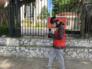 Bức ảnh chụp một người đàn ông đang đi bộ trên vỉa hè, bên cạnh một bức tường đá và một cổng sắt lớn. Người đàn ông đội mũ bảo hiểm, đeo khẩu trang, mặc áo khoác có mũ trùm đầu kẻ ca rô, bên ngoài là áo ghi lê phản quang màu cam, và đang chăm chú nhìn vào điện thoại di động. Trên cổng sắt có một tấm biển báo màu đỏ với dòng chữ có thể đọc được là "...VỰC CẤM TỤ...". Phía sau hàng rào, có thể thấy một biển báo cấm đỗ xe và một phần của một tòa nhà lớn có nhiều cửa sổ kính. Khung cảnh cho thấy đây có thể là khu vực bên ngoài một cơ quan hoặc một tòa nhà văn phòng.。（日本語訳）。写真には、石の壁と大きな鉄の門の横にある歩道を歩いている男性が写っている。男性はヘルメットをかぶり、マスクを着け、チェック柄のフード付きジャケットの上にオレンジ色の反射ベストを着用し、携帯電話を熱心に見つめている。鉄の門には、「...VỰC CẤM TỤ...」と読み取れる文字が書かれた赤い看板がある。この光景は、ここが何らかの機関またはオフィスビルの外のエリアである可能性を示している。