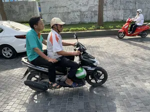 Bối cảnh là một con đường được lát gạch trong một khu vực đô thị. Hoạt động chính là người dân đang tham gia giao thông bằng xe máy. Ở trung tâm, hai người đàn ông đang đi trên một chiếc xe máy màu đen, trên thân xe có chữ 'AIR BLADE'. Người lái xe là một người đàn ông lớn tuổi, đội mũ bảo hiểm màu kem có họa tiết, đi dép lê. Người ngồi sau là một thanh niên mặc áo polo màu xanh ngọc. Phía trước xe máy có treo một chiếc mũ bảo hiểm màu xanh lá cây. Ở phía sau là một bức tường phù điêu lớn bằng đá, chạm khắc các hình ảnh mô tả nhiều người trong các hoạt động khác nhau, có thể là cảnh lịch sử hoặc lao động. Dọc theo bức tường có cây xanh và thảm cỏ. Bên trái có một phần của chiếc ô tô màu trắng. Ở phía xa bên phải, một người khác đang lái một chiếc xe máy tay ga màu đỏ.。（日本語訳）。背景は、都市部にあるレンガ敷きの道です。主な活動は、人々がバイクで交通に参加していることです。中央では、2人の男性が黒いバイクに乗っており、車体には「AIR BLADE」という文字があります。運転手は年配の男性で、模様の入ったクリーム色のヘルメットをかぶり、サンダルを履いています。後ろに乗っているのは、ターコイズブルーのポロシャツを着た若い男性です。バイクの前方には、緑色のヘルメットが掛けられています。後方には、石でできた大きなレリーフの壁があり、様々な活動に従事する多くの人々を描いた画像が彫られています。それは歴史的な場面か労働の場面かもしれません。壁に沿って緑の木々と芝生があります。左側には、白い車の一部が見えます。右の遠くでは、別の人が赤いスクーターを運転しています。