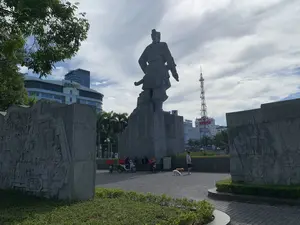 Bức ảnh chụp tại một quảng trường công cộng ở thành phố Thanh Hóa, có thể là Quảng trường Lam Sơn. Trung tâm của bức ảnh là một tượng đài bằng đá lớn, hoành tráng, khắc họa một nhân vật lịch sử, có thể là anh hùng Lê Lợi, trong tư thế oai phong với tay đặt trên chuôi gươm. Tượng đài được đặt trên một bệ đá cao. Hai bên là các bức tường phù điêu bằng đá lớn, chạm khắc các cảnh lịch sử. Xung quanh tượng đài là sân lát gạch và các bồn cây xanh được cắt tỉa gọn gàng. Trong hậu cảnh, có thể thấy các tòa nhà hiện đại, bao gồm một tòa nhà có logo "BIDV", một tòa nhà có chữ "vinaphone" và "ANH HOTEL", cùng với một tháp viễn thông cao. Về hoạt động, có một người đàn ông đang dắt hai con chó nhỏ đi dạo. Gần chân tượng đài, một nhóm người, có thể là tài xế giao hàng, đang ngồi nghỉ bên cạnh những chiếc xe máy của họ. Một trong những chiếc xe máy có gắn tấm biển quảng cáo với dòng chữ "THỢ ĐIỆN | SỬA CHỮA ĐIỆN NƯỚC" nhưng số điện thoại bên dưới không rõ.。（日本語訳）。この写真はタインホア市の公共広場で撮影されたもので、おそらくラムソン広場でしょう。写真の中心には、大きく壮大な石の記念碑があり、歴史上の人物、おそらく英雄レ・ロイが、剣の柄に手を置き威風堂々とした姿で描かれています。記念碑は高い石の台座の上に置かれています。両側には、歴史的な場面が彫刻された大きな石のレリーフの壁があります。記念碑の周りには、タイル敷きの広場と、きれいに刈り込まれた緑の植え込みがあります。背景には、「BIDV」のロゴがある建物、「vinaphone」と「ANH HOTEL」の文字がある建物、そして高い通信塔を含む現代的な建物が見えます。人々の活動については、一人の男性が2匹の小型犬を散歩させています。記念碑の足元の近くでは、一団の人々、おそらく配達ドライバーたちが、自分たちのバイクの隣で座って休んでいます。バイクの一台には「THỢ ĐIỆN | SỬA CHỮA ĐIỆN NƯỚC」（電気技師｜電気・水道修理）という文字が書かれた広告看板が取り付けられていますが、その下の電話番号ははっきりしません。