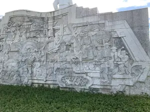 Bức ảnh chụp một bức phù điêu lớn bằng đá, là một phần của một khu tượng đài ngoài trời. Bức phù điêu tái hiện một cảnh tượng lịch sử, có thể là một cuộc diễu hành của quân đội hoặc một lễ hội. Nổi bật là hình ảnh một vị tướng lĩnh hoặc một vị vua đang cưỡi ngựa, được một người lính cầm lọng che. Theo sau là đoàn quân binh lính mặc quân phục cổ, đội nón và mang theo cờ hiệu. Một phần khác của bức phù điêu khắc họa cảnh người dân và binh lính đang múa rồng (hoặc lân) trong không khí tưng bừng, náo nhiệt. Phía sau có các công trình kiến trúc cổ. Phía trên cùng của bức tường có thể thấy phần chân của một bức tượng lớn. Dưới chân bức phù điêu là một thảm thực vật xanh tươi.。（日本語訳）。写真は、屋外の記念碑群の一部である、大きな石のレリーフを写している。レリーフは、軍隊のパレードか祭りである可能性のある、歴史的な光景を再現している。際立っているのは、馬に乗り、兵士に日傘を差してもらっている将軍または王の姿である。後ろには、古い軍服を着て笠をかぶり、旗印を掲げた兵士の一団が続いている。レリーフの別の部分は、人々や兵士が賑やかで活気のある雰囲気の中で龍舞（または獅子舞）を踊っている光景を描写している。後ろには古い建築物がある。レリーフの足元には、青々とした植生が広がっている。