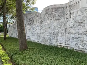 Bức ảnh chụp một bức tường phù điêu bằng đá lớn, uốn cong, đặt trong một không gian công cộng như công viên hoặc quảng trường. Bức tường được chạm khắc công phu với nhiều hình ảnh mô tả các sự kiện lịch sử hoặc các cảnh sinh hoạt văn hóa của Việt Nam. Trên bức phù điêu, có thể nhận thấy hình ảnh của nhiều người trong trang phục truyền thống, binh lính, người dân đội nón lá, các công trình kiến trúc cổ như cổng thành, và các sinh vật thần thoại như rồng hoặc lân. Các hoạt động được khắc họa bao gồm các đoàn diễu hành, trận chiến và cuộc sống thường ngày. Xung quanh chân tường là thảm cây xanh. Phía trước có những thân cây to. Ở phía sau, một tòa nhà cao tầng hiện đại với mặt tiền bằng kính và một logo màu vàng có thể được nhìn thấy. Không có văn bản nào có thể đọc được trong ảnh.。（日本語訳）。写真は、公園や広場のような公共空間に設置された、湾曲した大きな石のレリーフの壁を写しています。壁には、ベトナムの歴史的な出来事や文化的な生活風景を描写した多くの図像が精巧に彫刻されています。レリーフの上には、伝統的な衣装をまとった多くの人々、兵士、ノンラーをかぶった人々、城門のような古代の建築物、そして龍や麒麟のような神話上の生き物の姿が見て取れます。描かれている活動には、パレードの行列、戦闘、そして日常生活が含まれます。壁の根元の周りには、緑の植物が広がっています。手前には太い木の幹があります。後方には、ガラス張りのファサードと黄色のロゴを持つ近代的な高層ビルが見えます。写真の中には、読み取れる文字はありません。