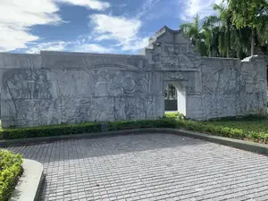 Bức ảnh chụp một bức tường phù điêu lớn bằng đá, nằm ở một không gian công cộng ngoài trời như công viên hoặc quảng trường. Bức tường có một cổng vòm ở trung tâm. Bề mặt tường được chạm khắc công phu, tái hiện các cảnh sinh hoạt, lao động và các sự kiện lịch sử. Các hình chạm khắc bao gồm hình ảnh con người trong trang phục truyền thống, thuyền buồm trên biển, những người thợ đang làm việc, các học giả, và biểu tượng âm dương. Không có văn tự hay chữ viết nào có thể nhận dạng được trên bức phù điêu. Phía trước bức tường là một khoảng sân lát đá. Dọc chân tường được trang trí bằng các hàng cây bụi cắt tỉa gọn gàng. Phía sau và xung quanh là những cây cọ cao lớn. Bầu trời trong xanh, có mây trắng.。（日本語訳）。写真は、公園や広場のような屋外の公共スペースにある、大きな石のレリーフ壁を写しています。壁の中央にはアーチ型の門があります。壁の表面は精巧に彫刻されており、生活や労働の場面、そして歴史的な出来事を再現しています。彫刻には、伝統的な衣装を着た人々の姿、海上の帆船、働く職人たち、学者たち、そして陰陽のシンボルが含まれています。レリーフの上には、識別できる文字や文章はありません。壁の前には石畳の中庭があります。壁の根元に沿って、きれいに刈り込まれた低木の列で飾られています。後ろと周りには高いヤシの木があります。空は青く澄み、白い雲があります。